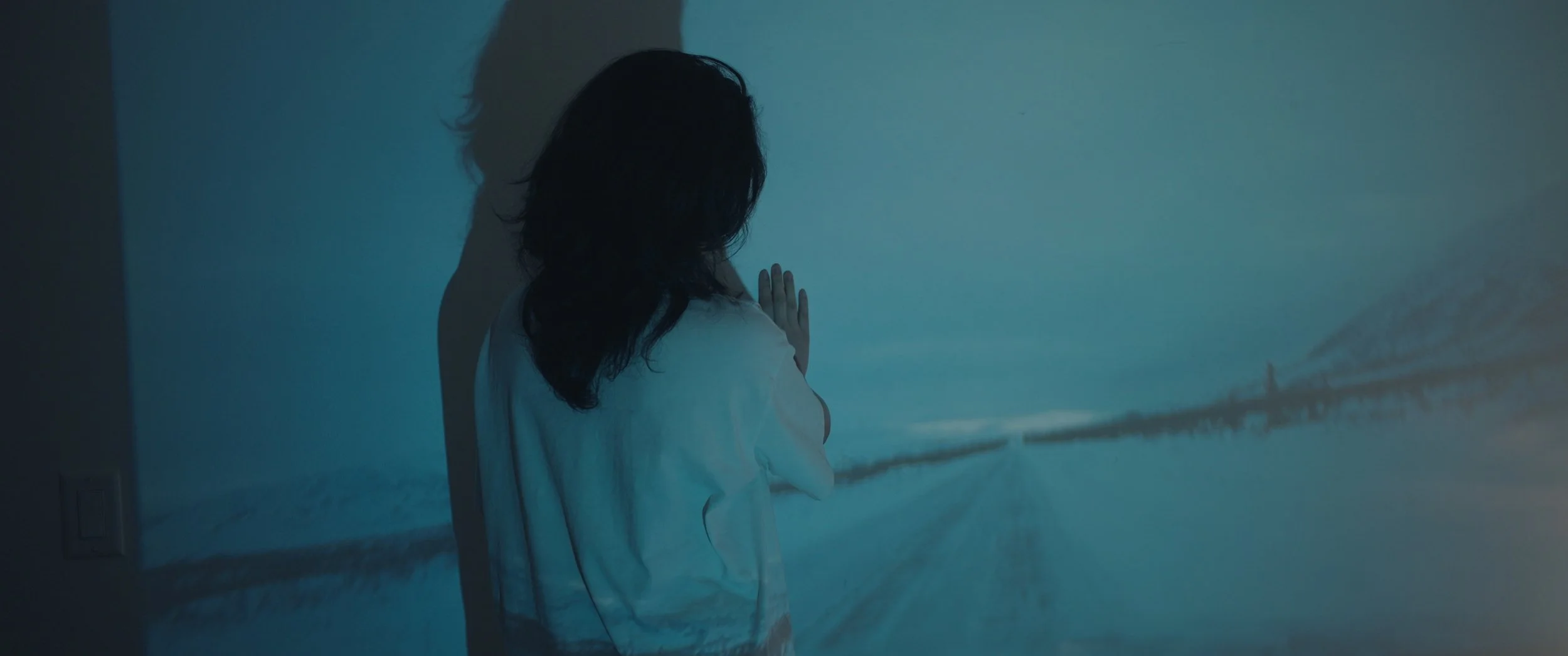 A person with long dark hair facing a wall with a painting of a bridge over water, with hands pressed against the wall.