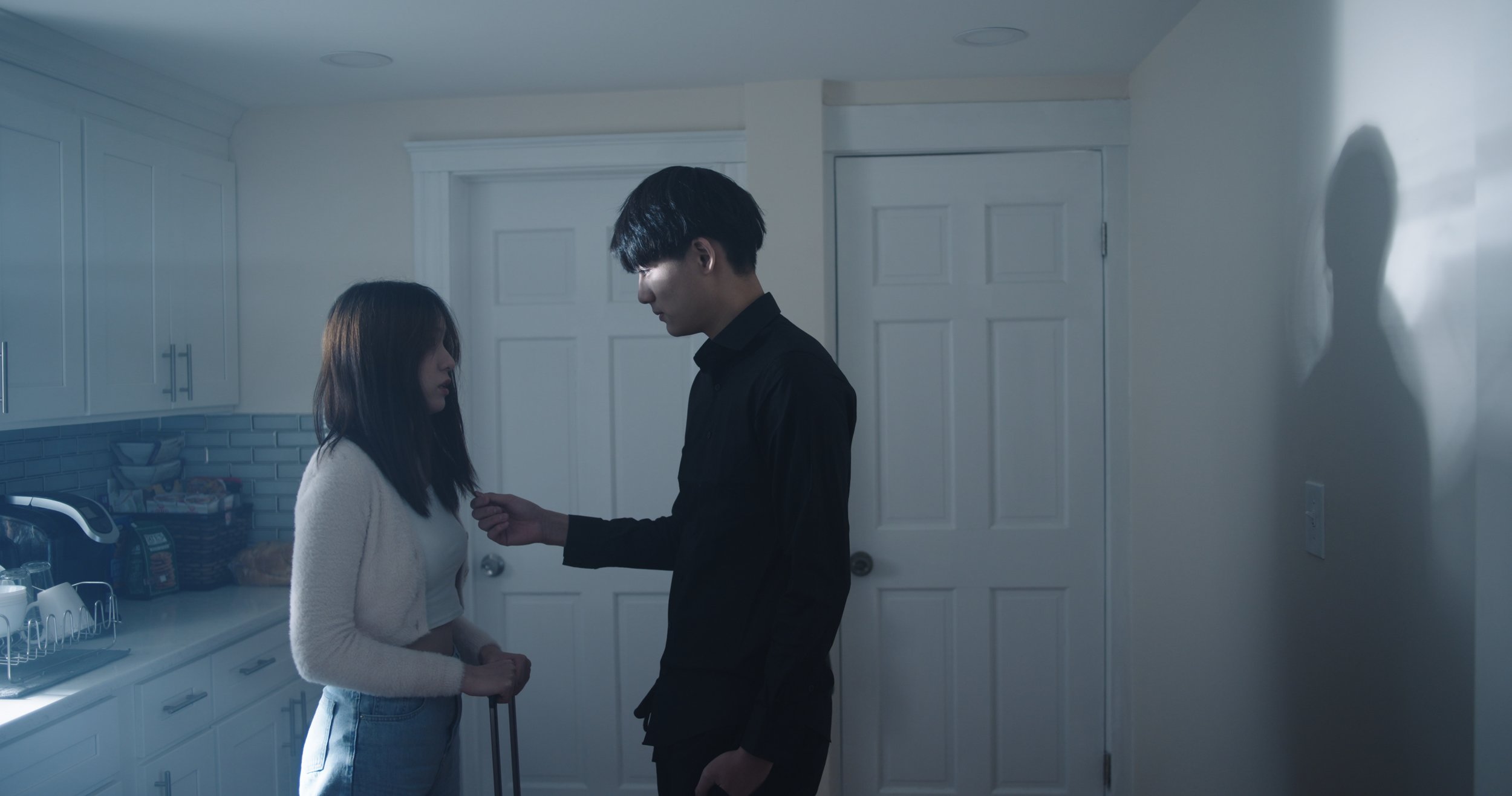 A man in a black shirt and a woman in a white sweater standing close in a kitchen, with the man appearing to check or adjust the woman's collar, while she looks down.
