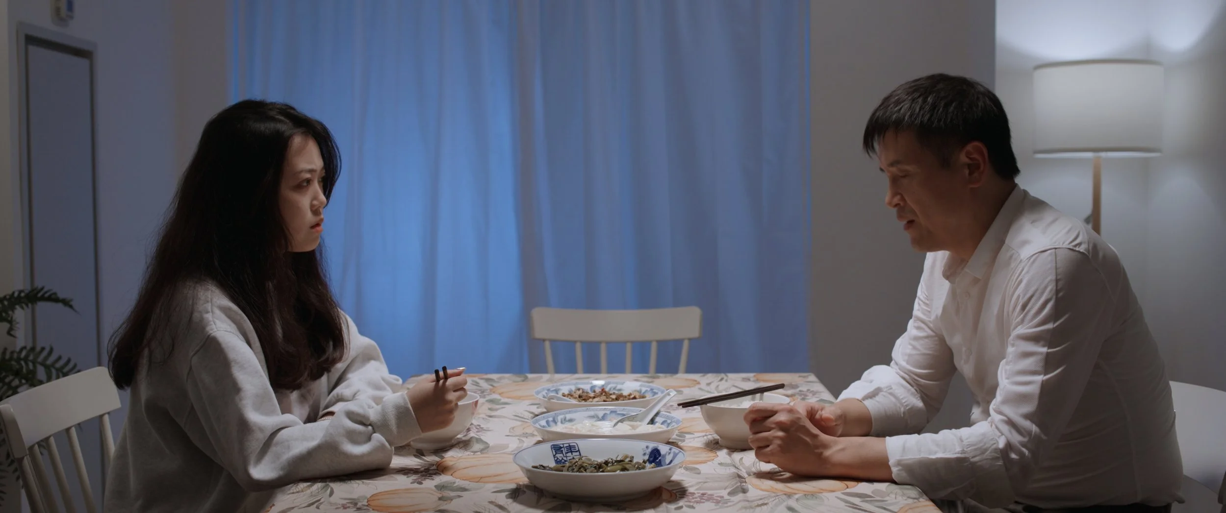A young woman and an older man sitting at a dining table, facing each other with tense expressions. The table has several bowls of food, and the background includes a lamp and a blue curtain.
