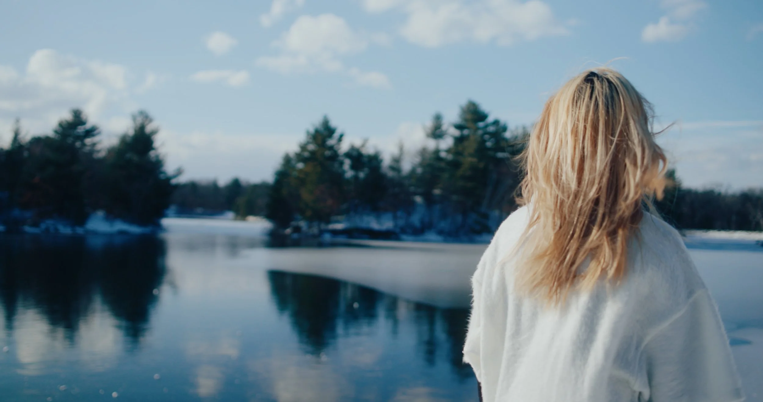 A woman with long blonde hair standing near a body of water with trees in the background, during a cloudy day.