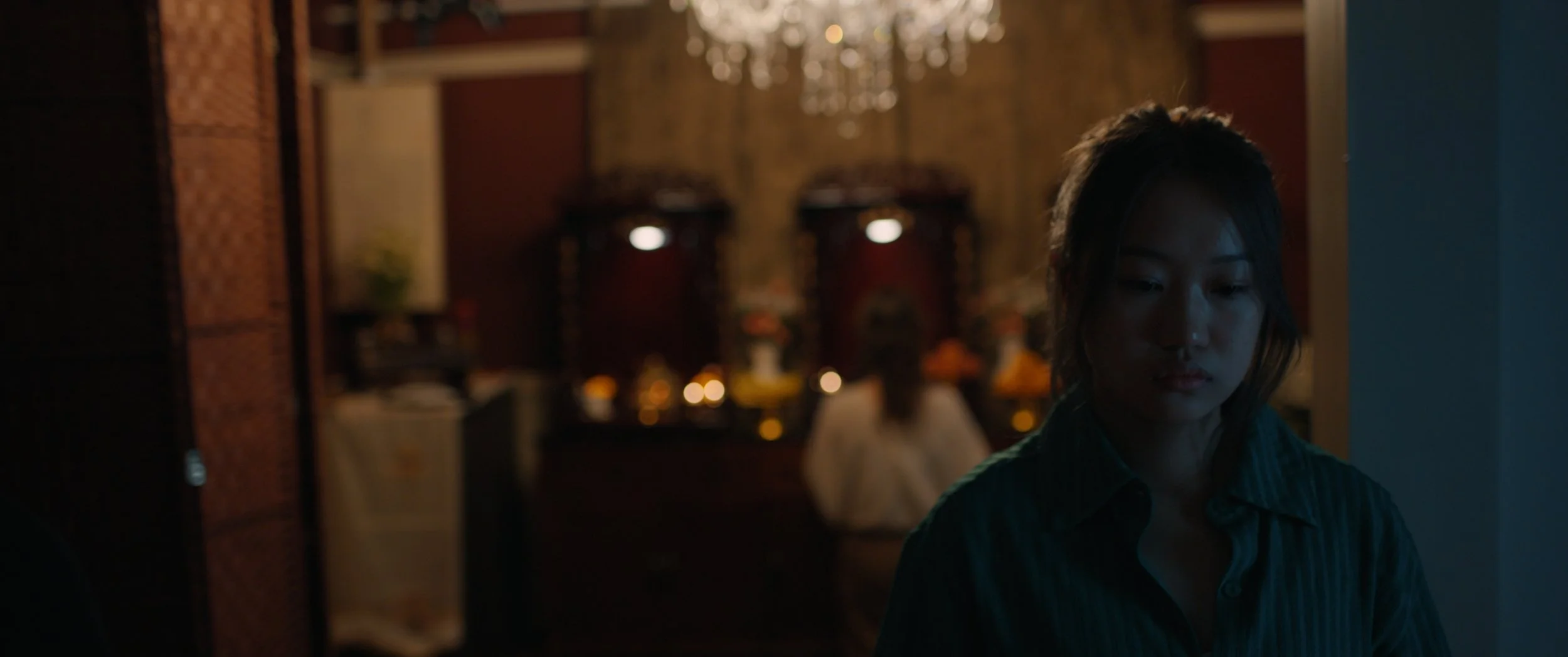 A woman with short hair looking down in a dimly lit room with blurred background of a chandelier and decorations.