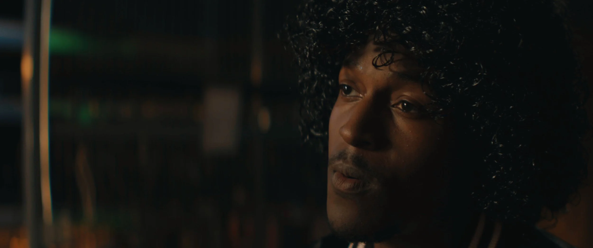 Close-up of a man with curly hair looking thoughtfully at computer screens in a dimly lit environment.