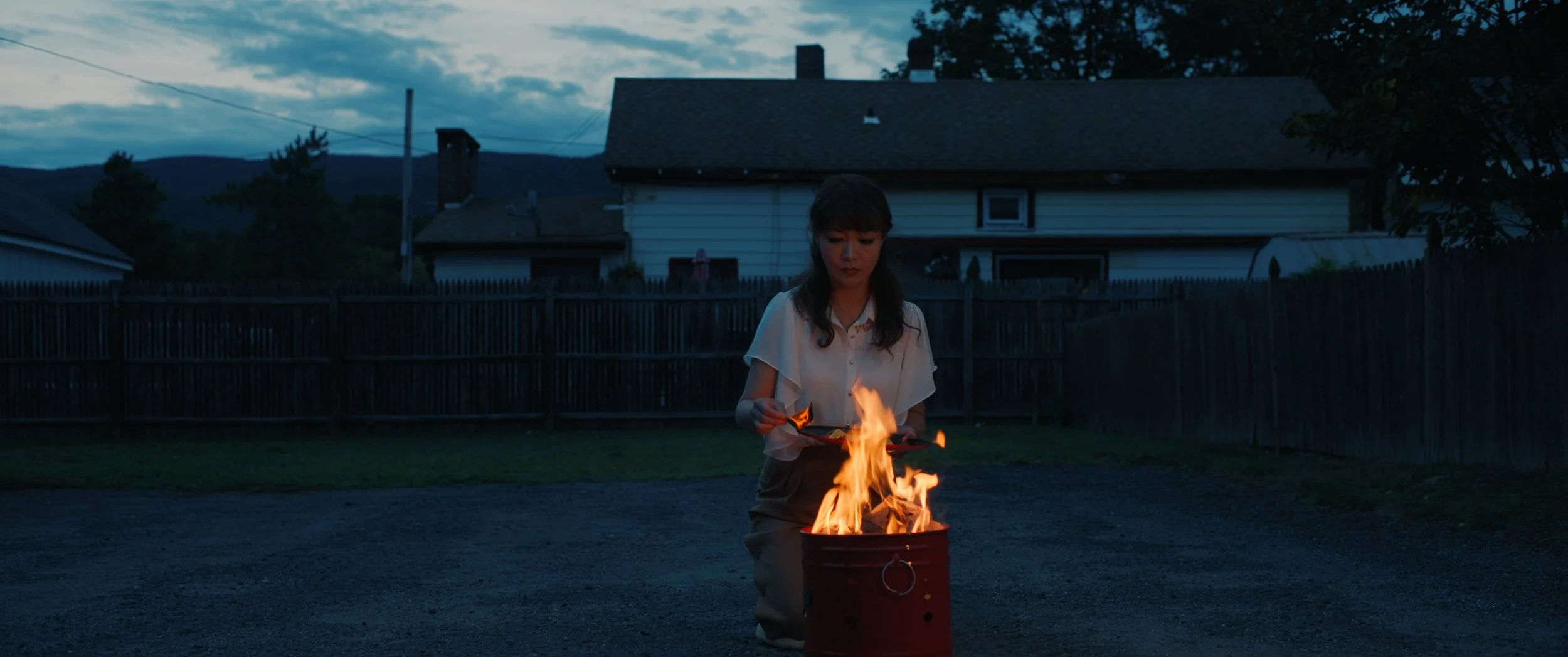 A woman standing outdoors during dusk, holding a burning object over a red barrel with flames.