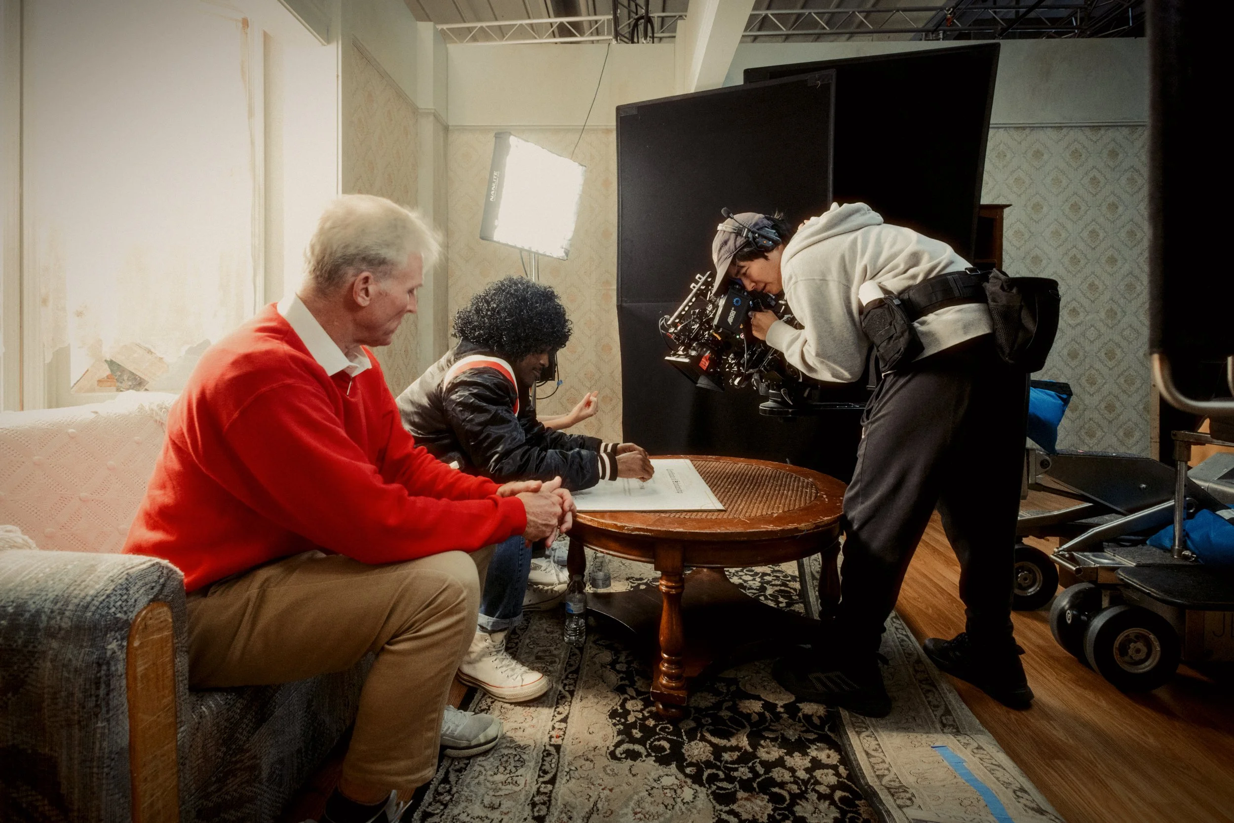 Film crew members setting up on a set with a camera and lighting equipment, preparing for a scene with actors seated on a couch and a table.