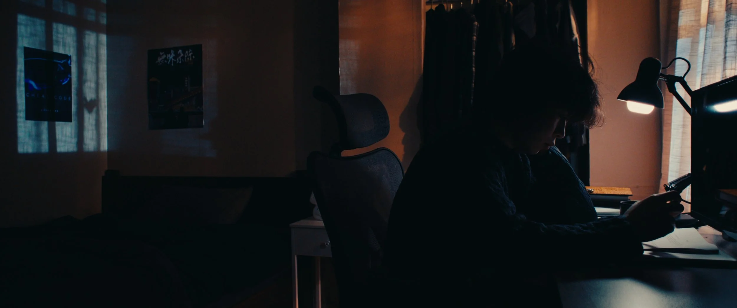 A person with dark hair sitting at a desk in a dimly lit room, looking at a phone, with a desk lamp illuminating the workspace and a closet with hanging clothes in the background.