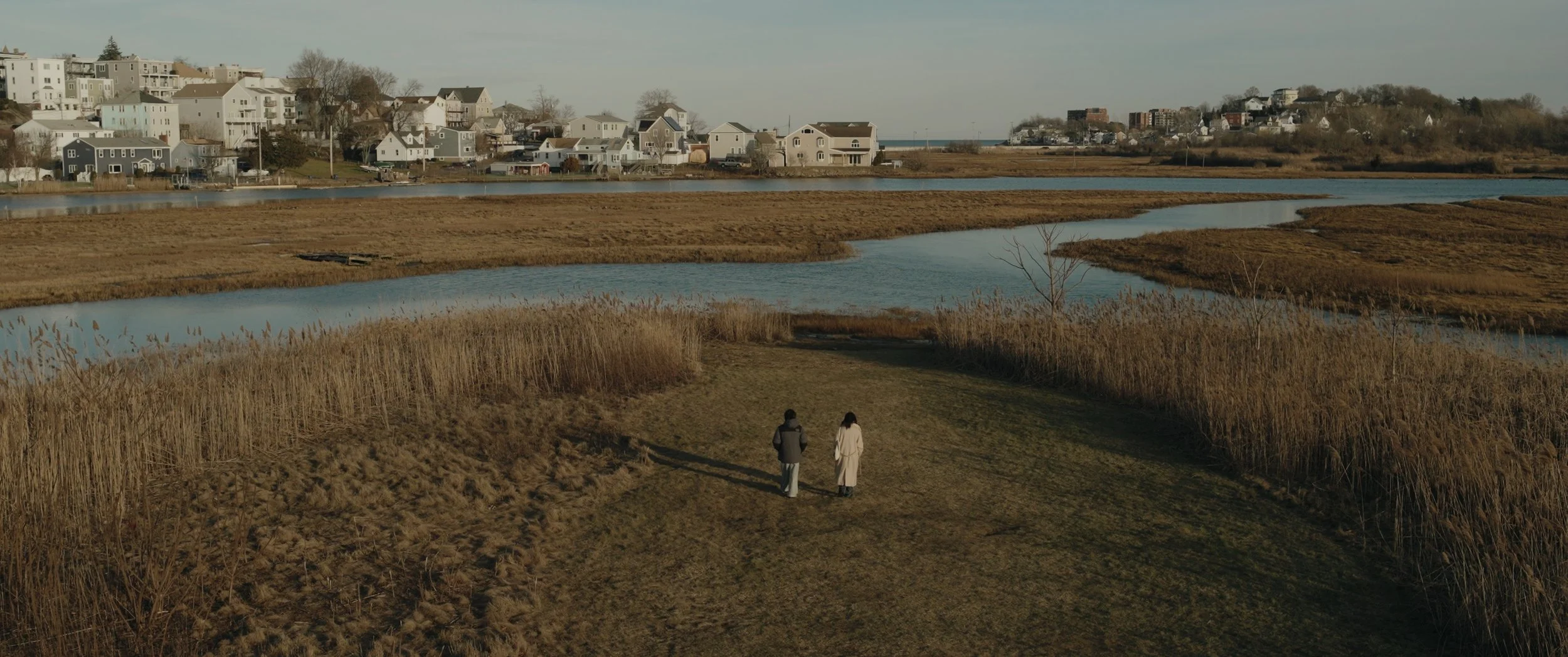 Two people walking on a grassy trail near a marsh with water and tall grass, with a residential neighborhood of houses in the background.
