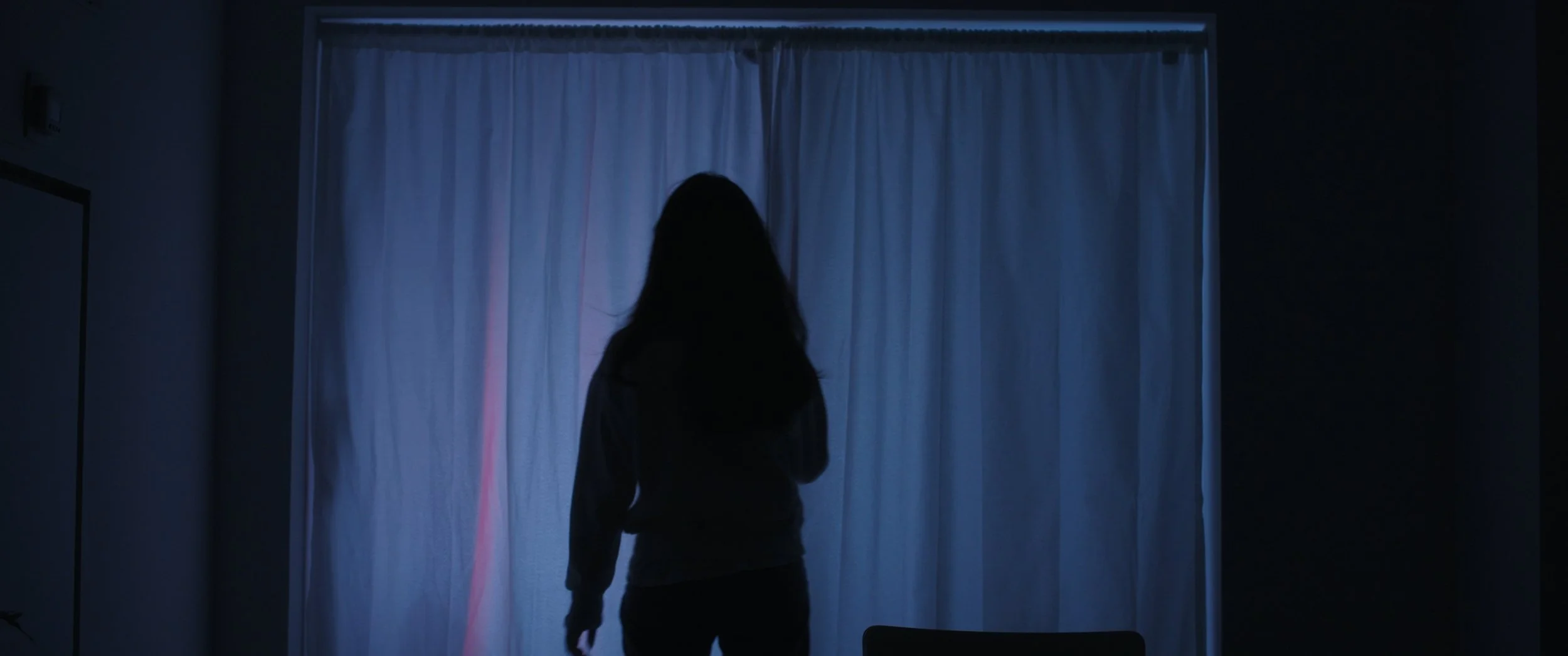 A person with long hair standing indoors in front of closed curtains, illuminated by blue light from outside, creating a silhouette.
