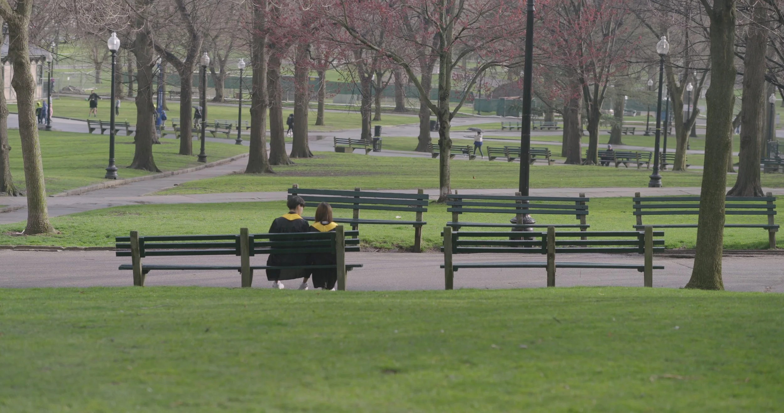 Two individuals sitting on a park bench, wearing black robes with yellow accents, surrounded by green grass and trees, with walking paths and other park visitors in the background during daytime.