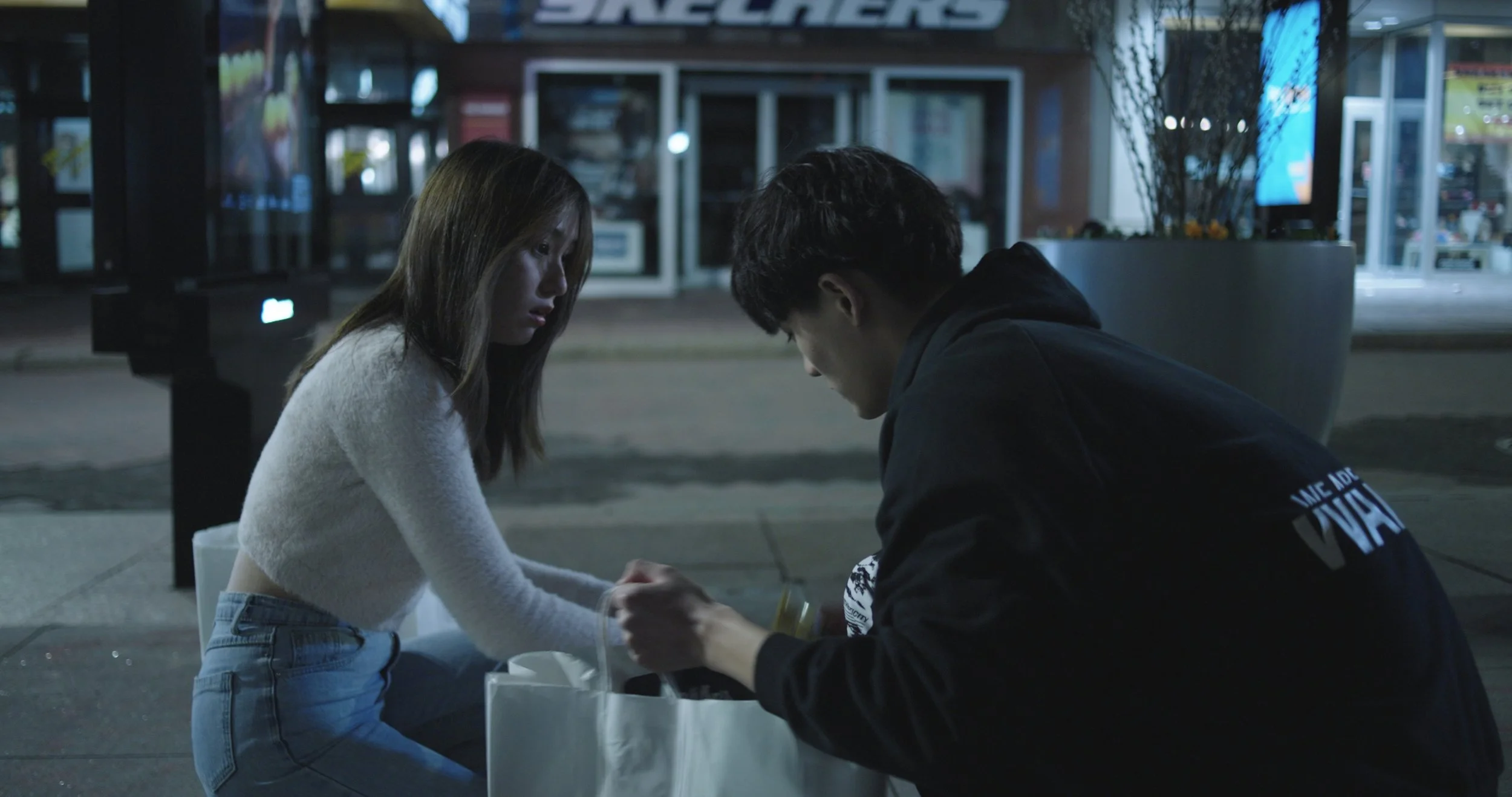 A young woman and a young man are sitting at a table on a city sidewalk at night, holding hands and looking at each other. The woman has long hair and is wearing a light-colored sweater and jeans, while the man is wearing a dark hoodie. The background shows a storefront with glass doors and windows, lit from inside, and a large planter with tall plants nearby.