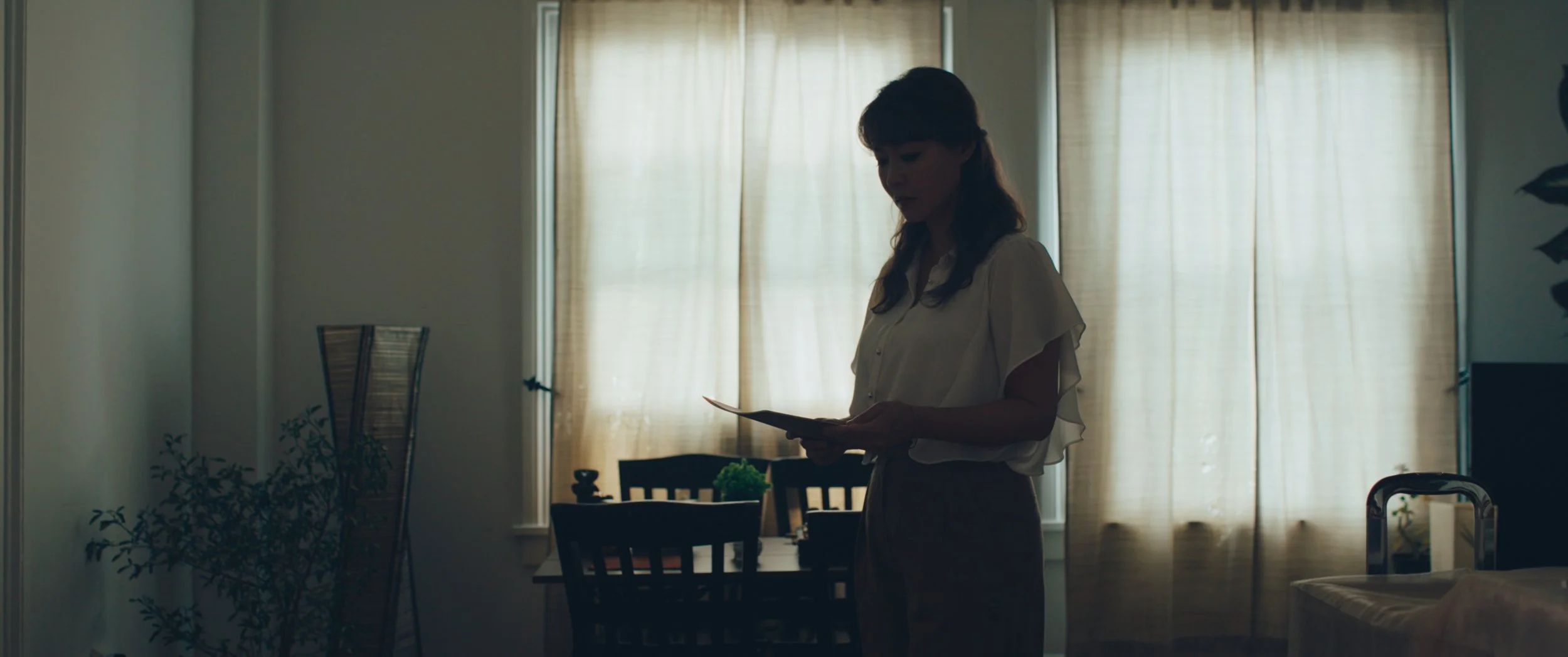 A woman stands in a dining room with beige curtains and a table with chairs, looking at a tablet in her hands.