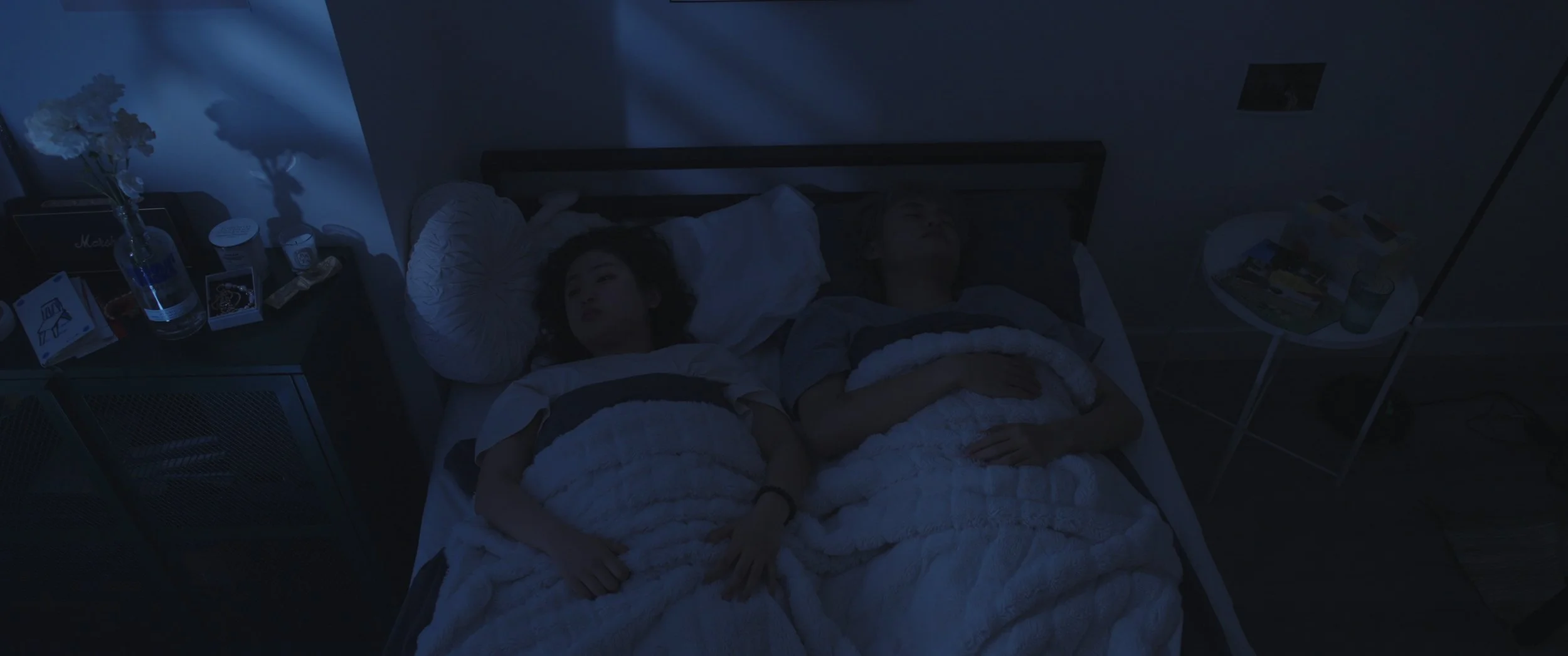 Two women lying in bed at night, peacefully sleeping, in a dimly lit room with a side table holding a water bottle, flowers, and various items.