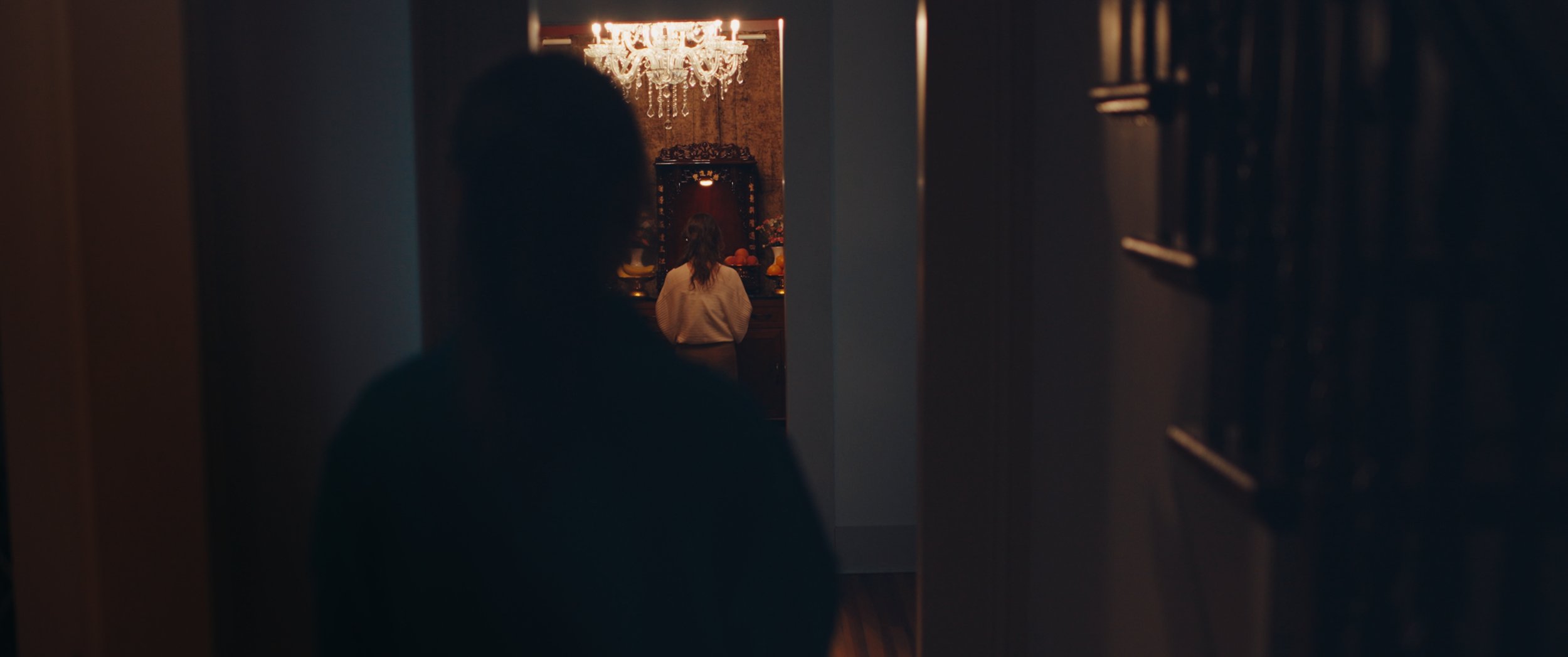 A person standing in a doorway looking into a dimly lit room with an ornate altar, flowers, candles, and a chandelier.