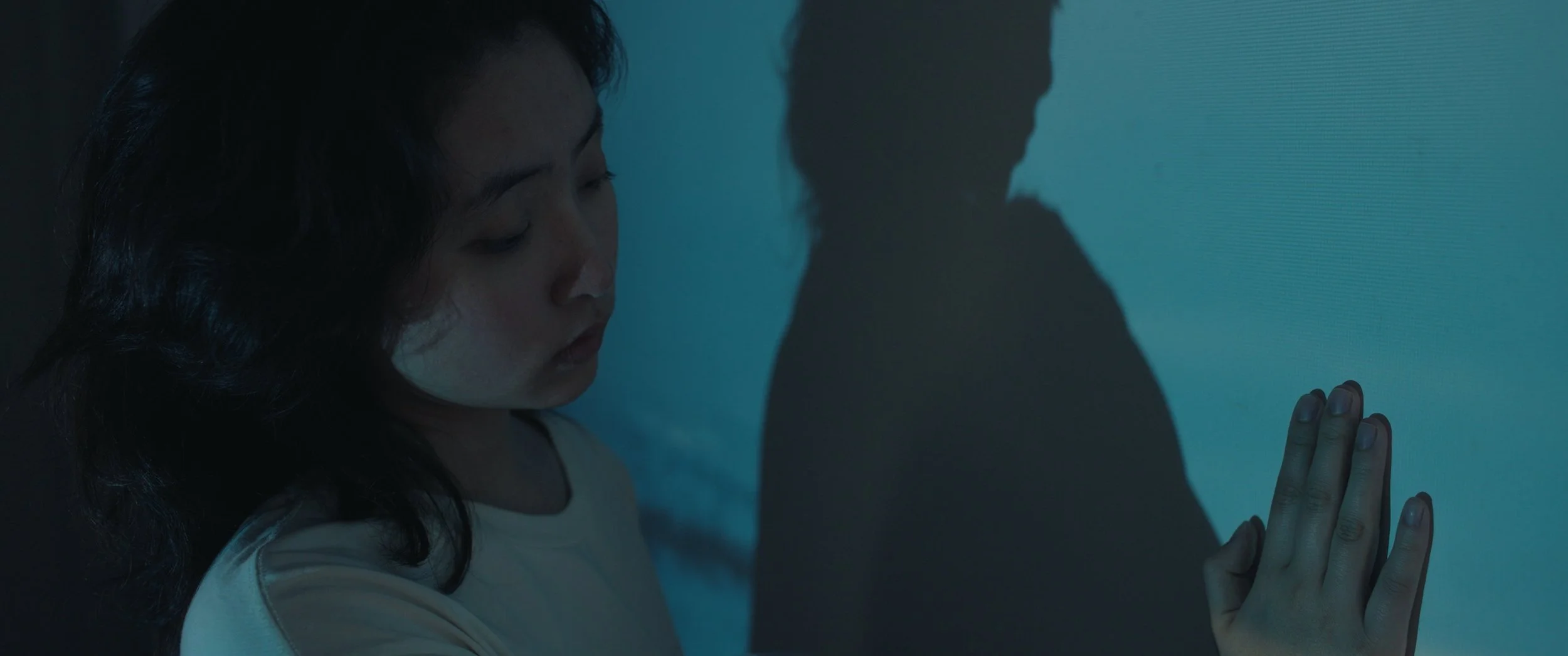 A young woman with dark, curly hair touching a textured blue wall with her hand, casting a shadow on the wall