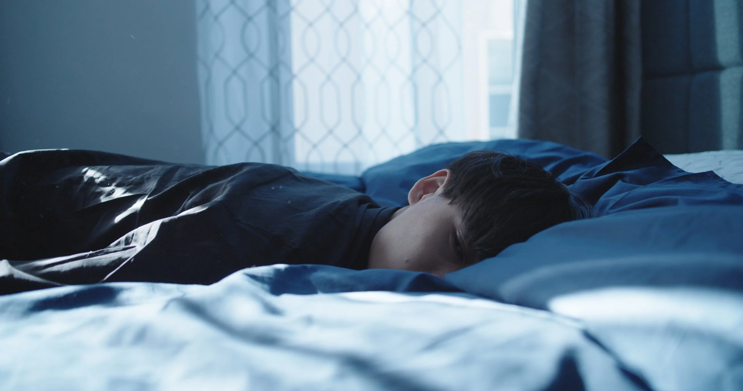 A person with short dark hair lying face down on a bed with blue bedding, in a room with natural light coming through a window with patterned curtains.