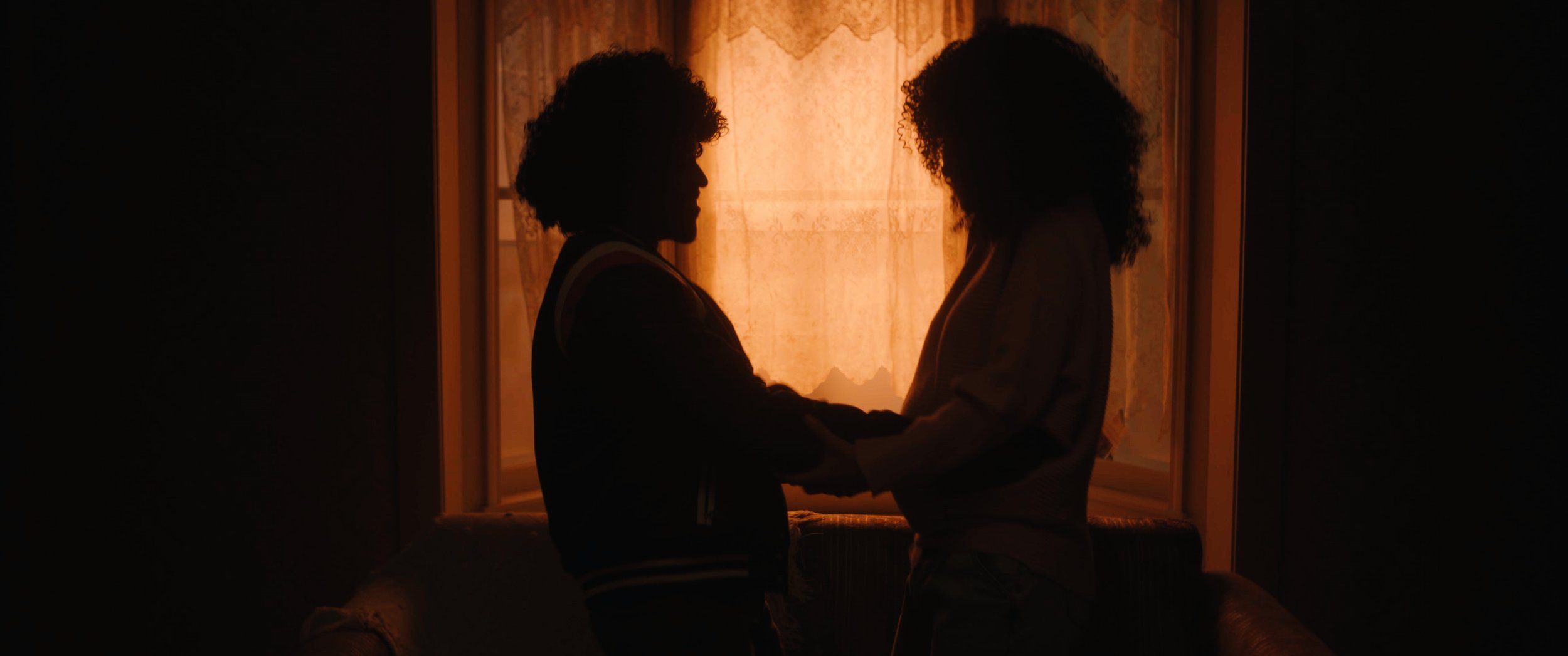 Two people with curly hair holding hands and facing each other in front of a window with a curtain, backlit by warm orange light, creating silhouettes.