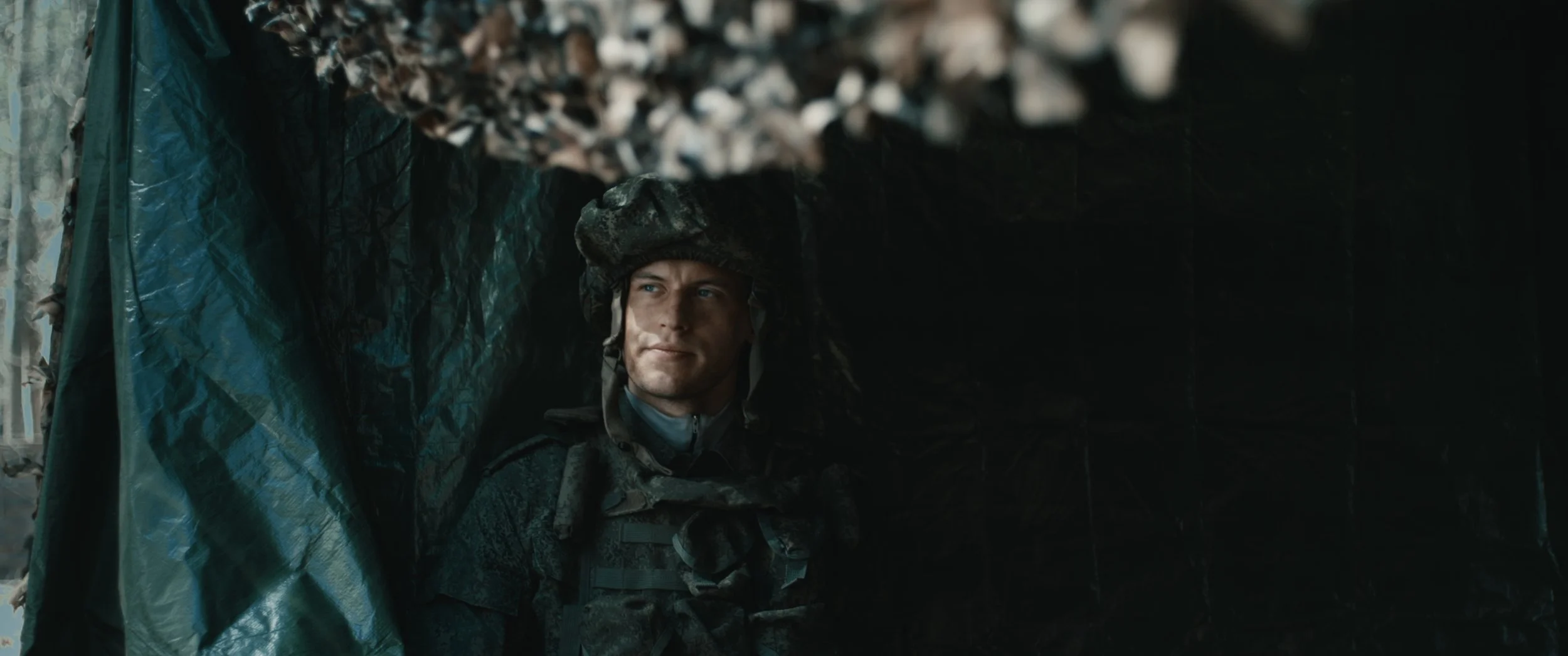 A soldier in camouflage uniform and helmet looking calmly from a hidden position within a small dark opening, possibly a tent or dugout.
