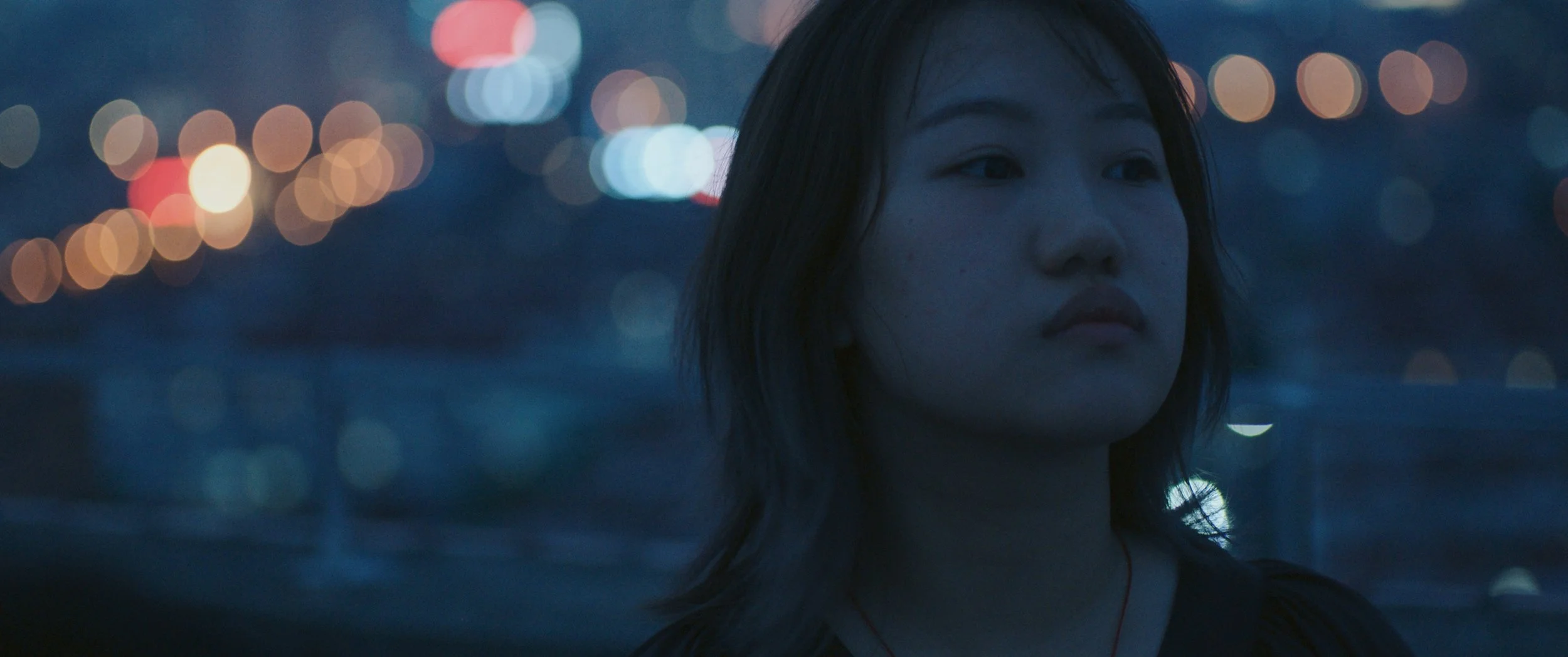 A young woman with dark hair looks into the distance at dusk with city lights blurred in the background.