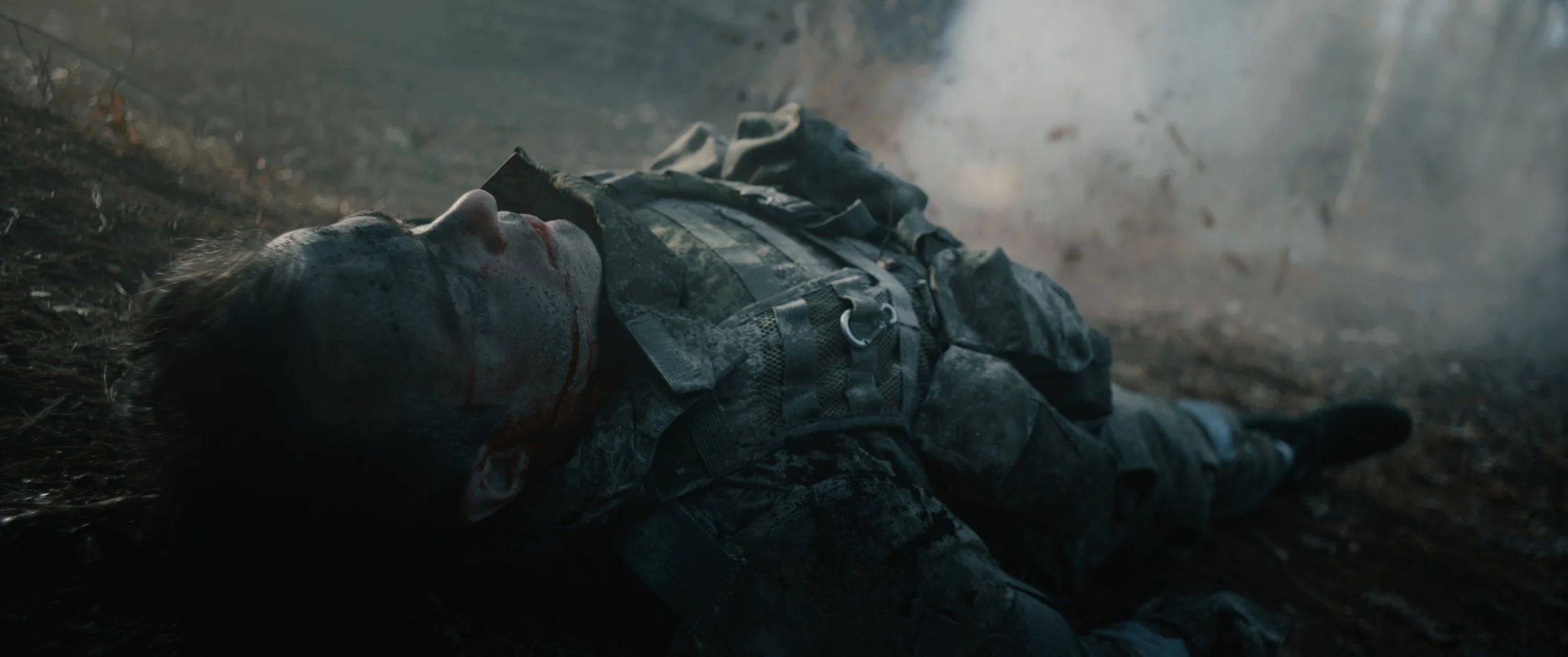 A soldier lying on the ground in a forest, wearing camouflage uniform and gear, with blood on his face and background smoke.