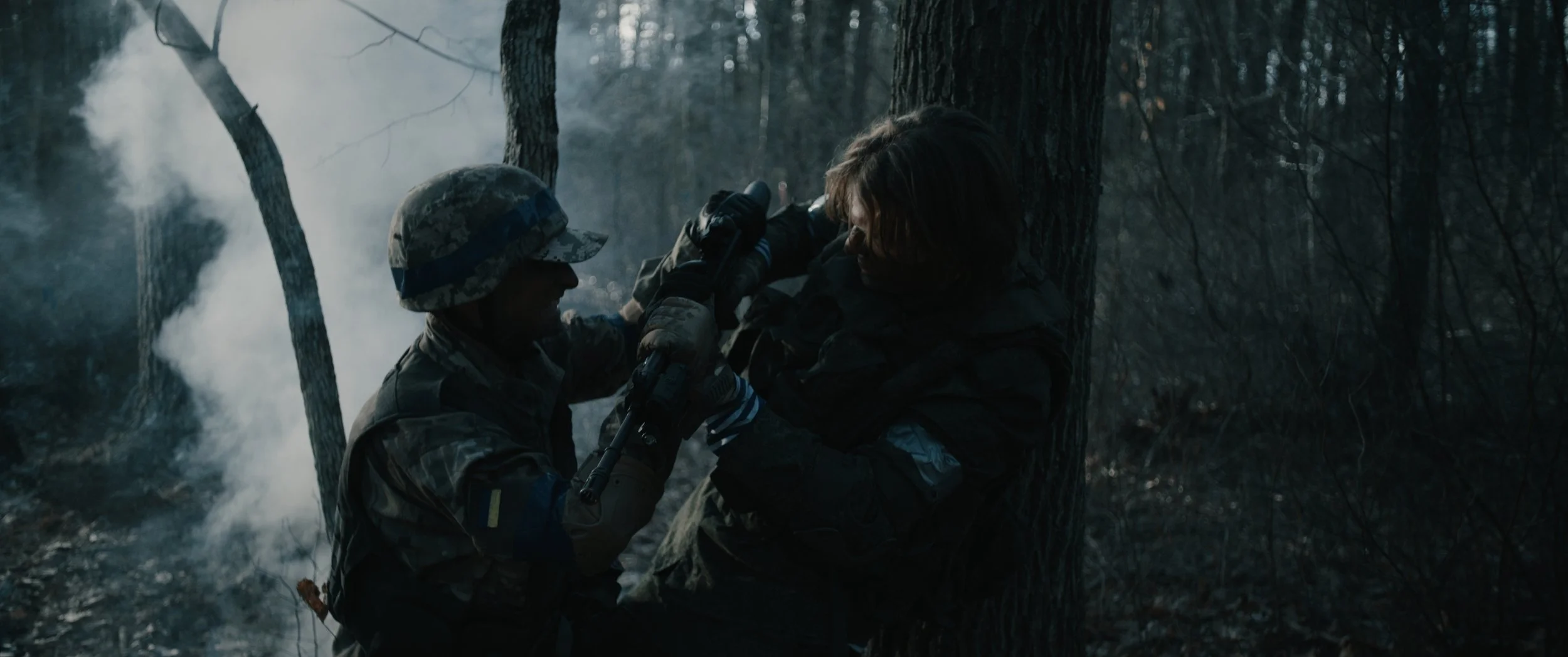 A soldier in camouflage attire attempting to rescue another soldier with brown hair who is leaning against a tree in a dark, smoky forest.