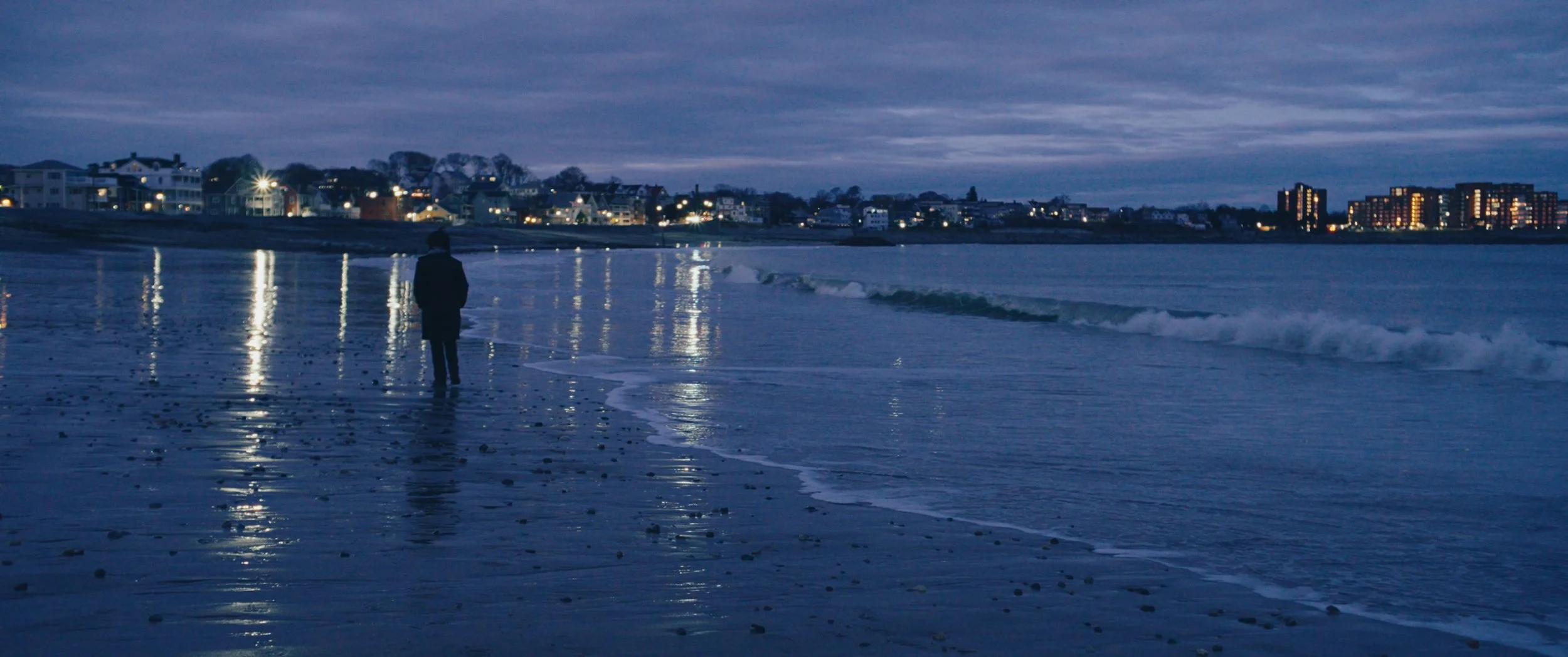 A person walking on the beach at dusk or dawn, with the shoreline, waves, and reflections of city lights on the water, and a cityscape with buildings in the distance.