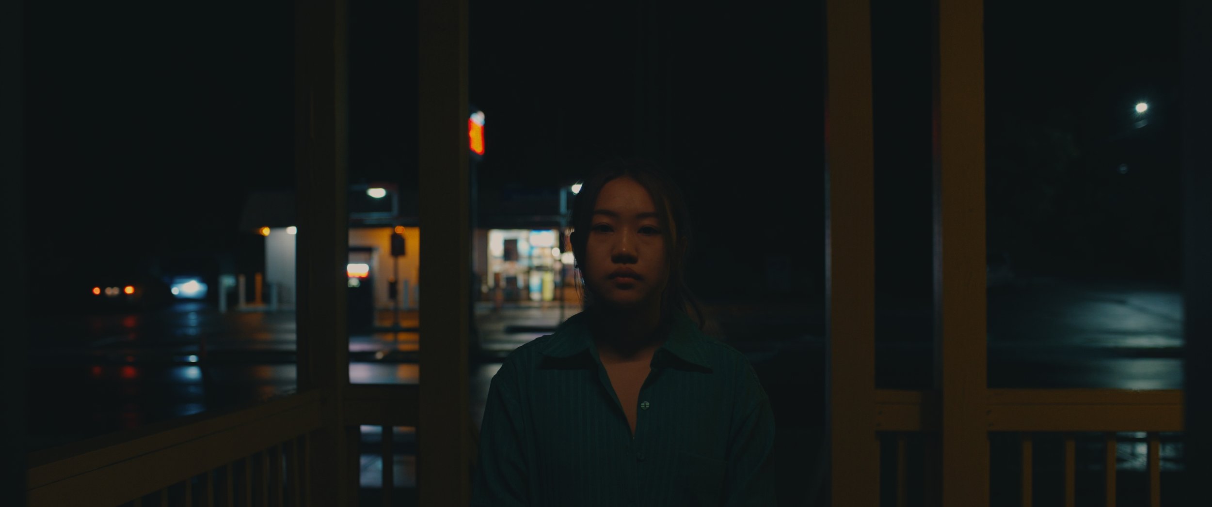 A young woman standing at night near a bus stop shelter, illuminated by distant city lights and a nearby convenience store.