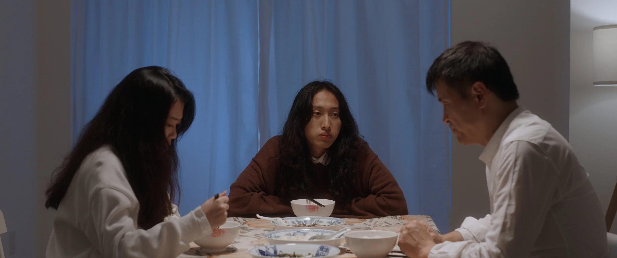 Three people sitting at a dining table with bowls and dishes, one woman in a brown sweater looking tired or upset, the others in white shirts focused on the table, blue curtains in the background.