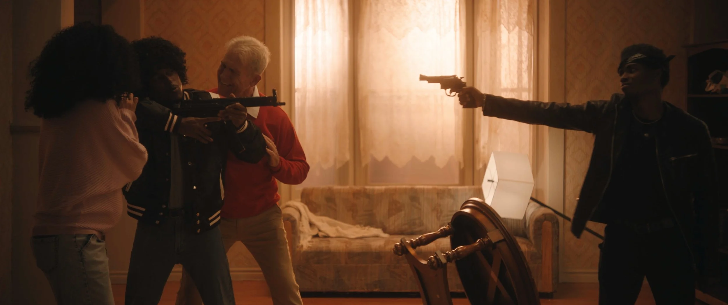 Two young women and two men holding guns in a tense standoff inside a living room, with a broken table on the floor.