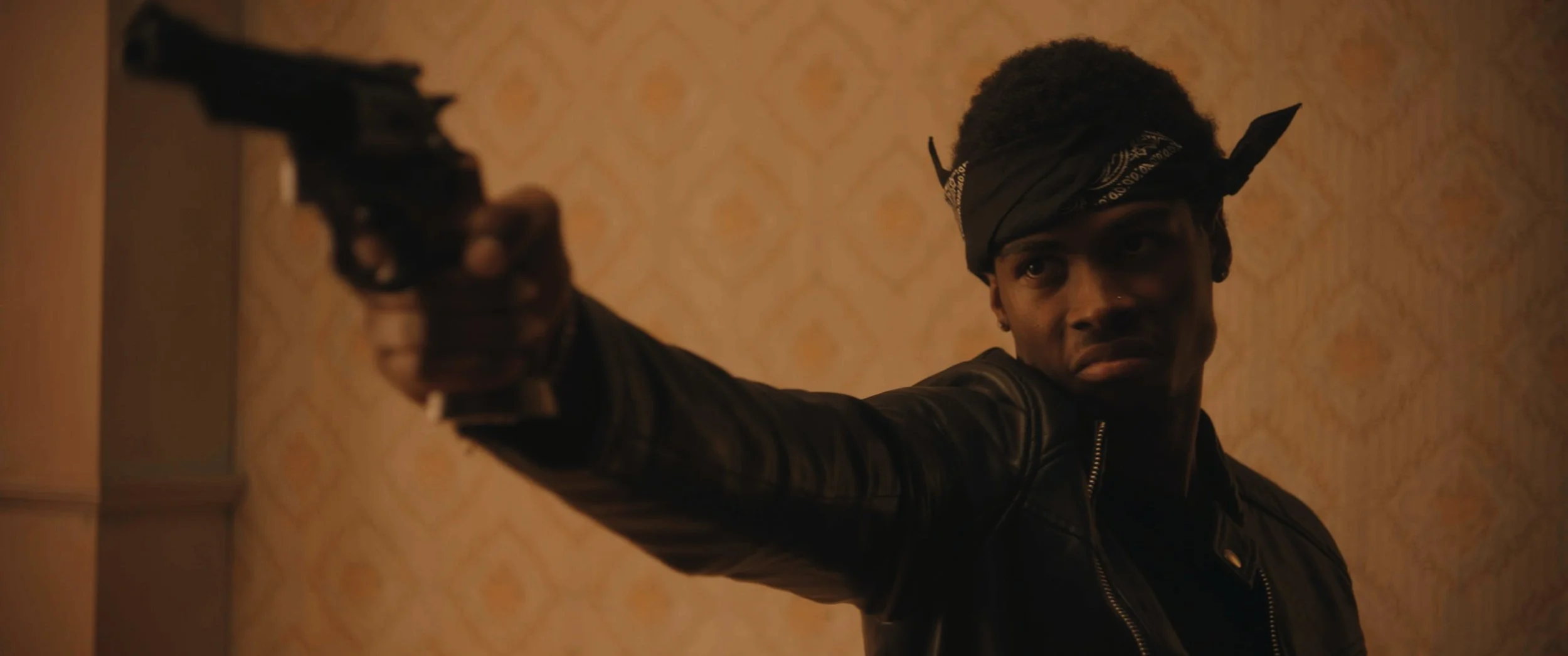 A person with short hair, wearing a black bandana on their head and a black leather jacket, aiming a handgun with an intense expression inside a room with beige patterned wallpaper.