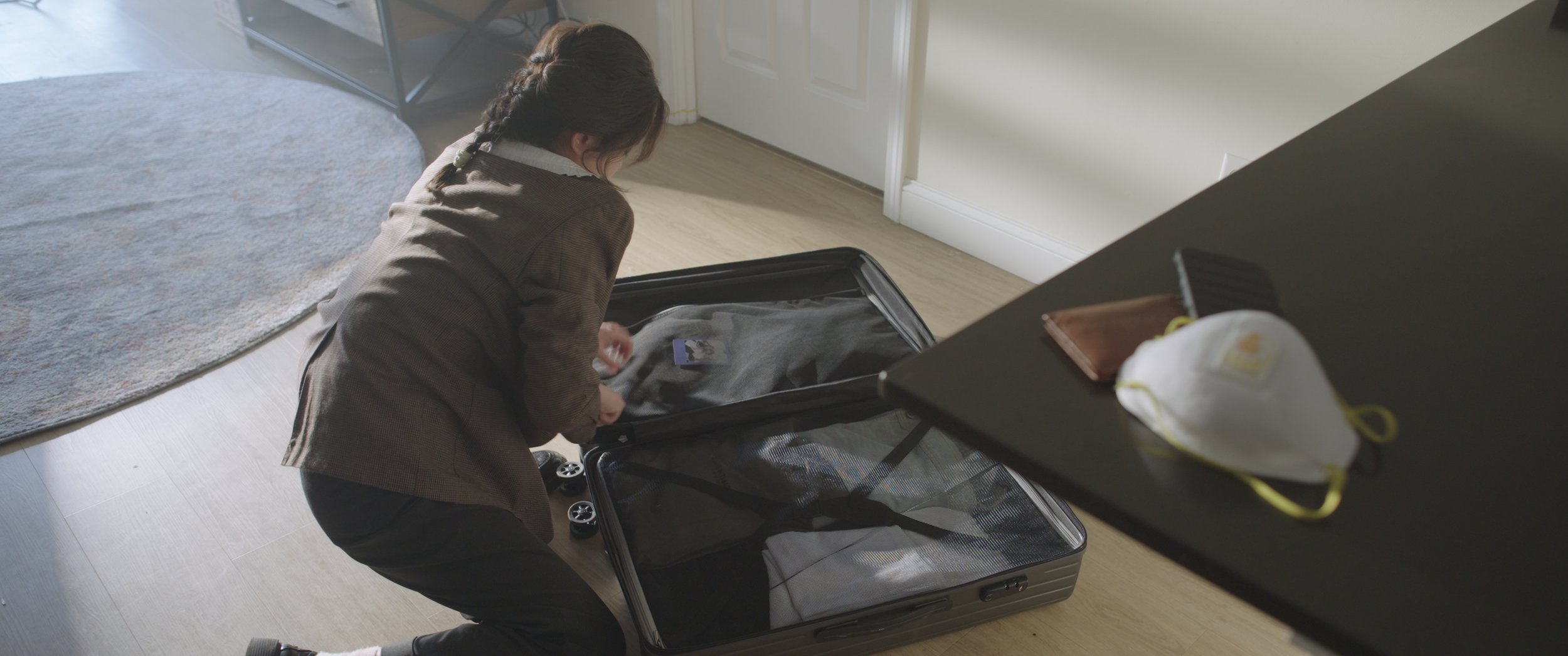 A woman packing a suitcase in a room with a door and a rug, with face mask and small electronic device on a nearby table.
