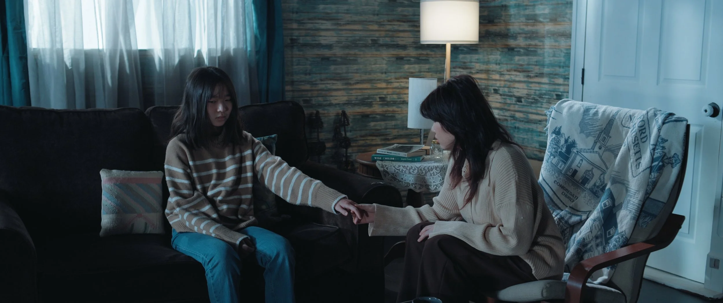 Two women sitting in a cozy room, holding hands with a dark sofa, a small side table with books, a nightstand lamp, and a wooden wall in the background.