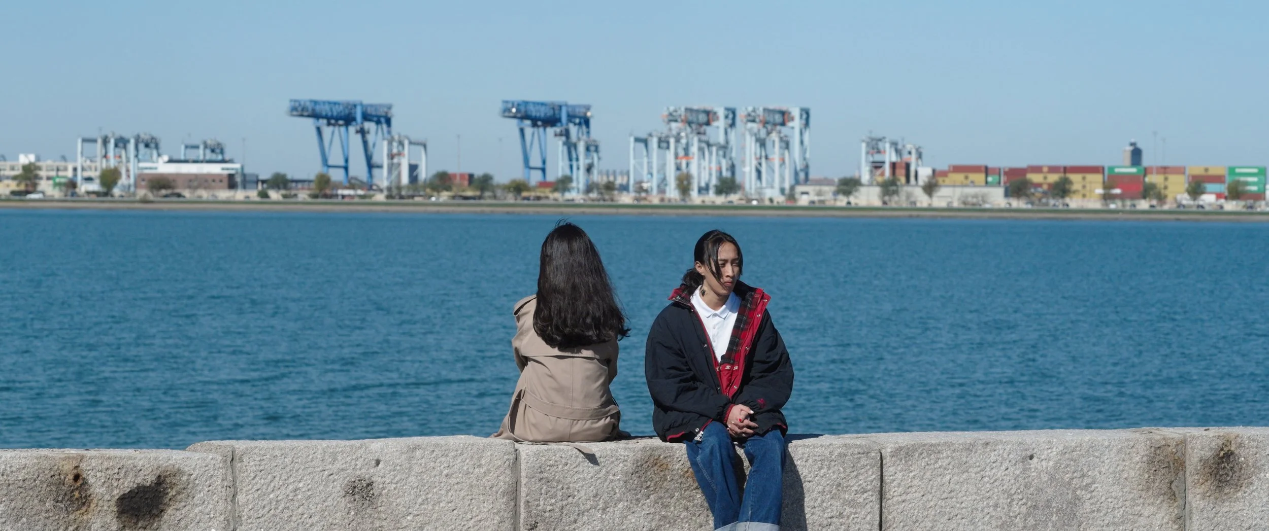 Two women sitting on a stone ledge by the water with an industrial port and cranes in the background.