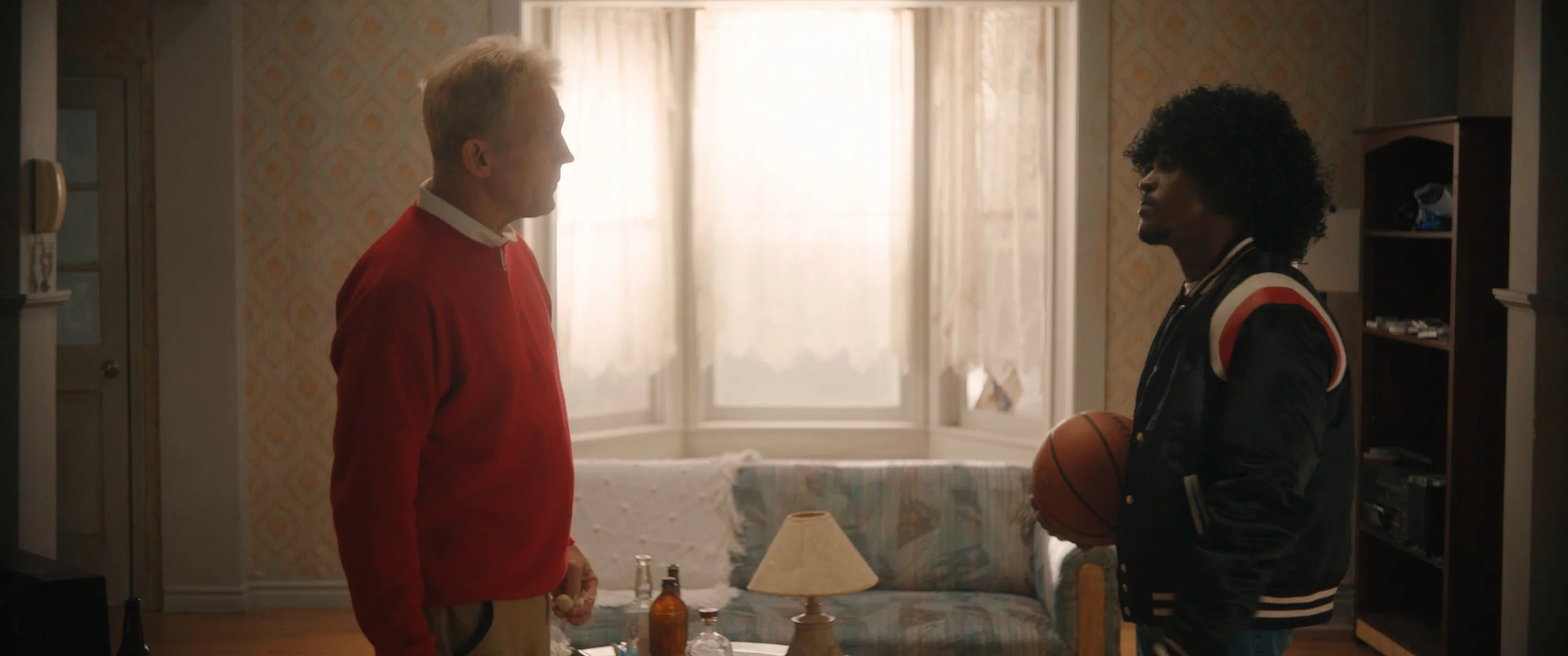 Two men standing face-to-face in a living room, one holding a basketball, with a table and bottles in front of them.