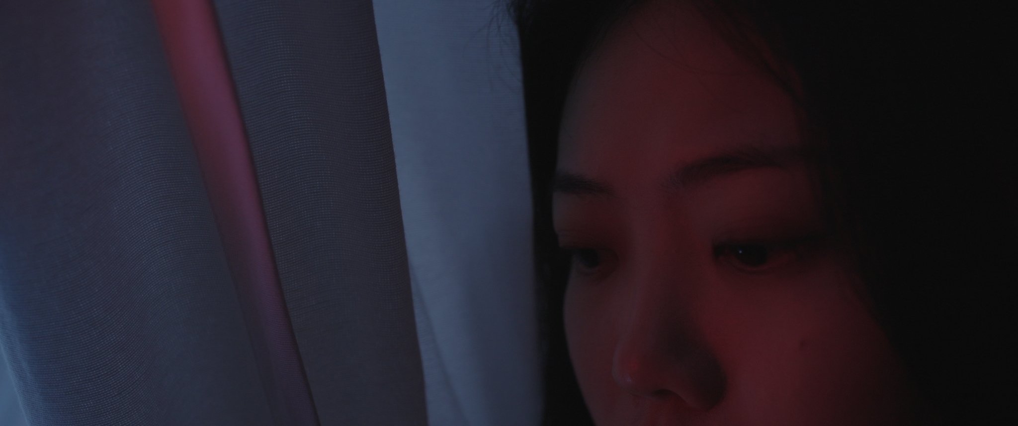 Close-up of a young woman’s face as she looks through a window or screen, with gentle lighting framing her face.