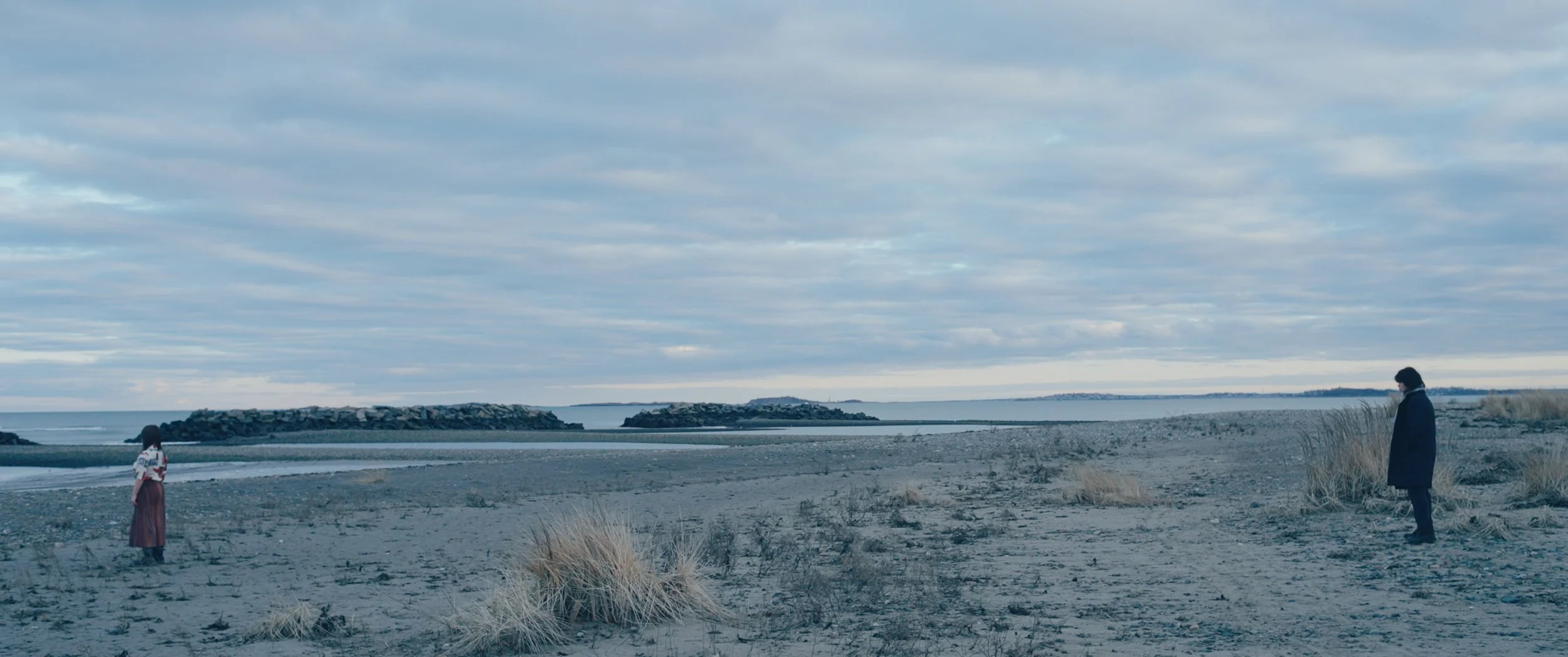 A person in a long coat and a woman in a long skirt standing on a sandy beach near the water, with grassy dunes in the foreground and rocky breakwaters in the background under an overcast sky.
