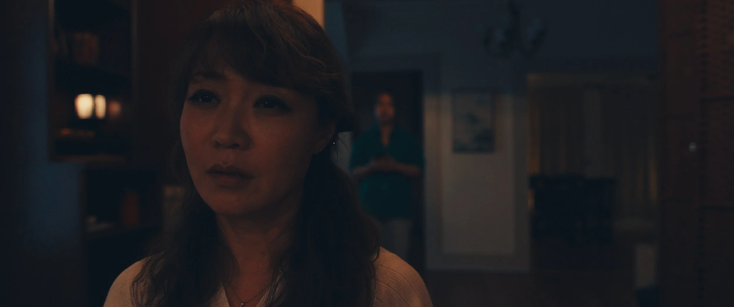 A woman with brown hair and a light-colored top is in focus, with a slightly emotional expression on her face. In the background, a person in a teal shirt stands in a dimly lit room, near a doorway.