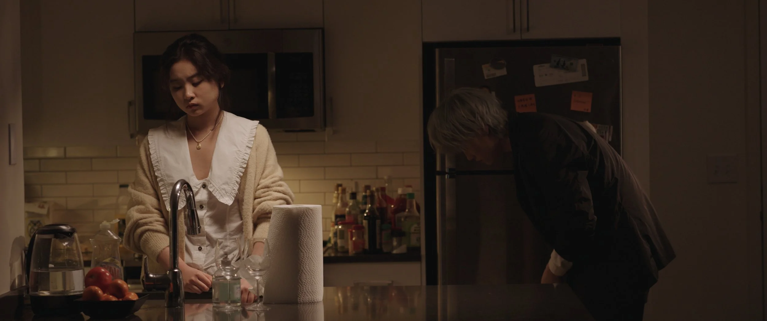 A woman with short dark hair standing at a kitchen counter, looking down, wearing a beige cardigan over a white top, with a man with gray hair bending down beside her in a dimly lit kitchen.