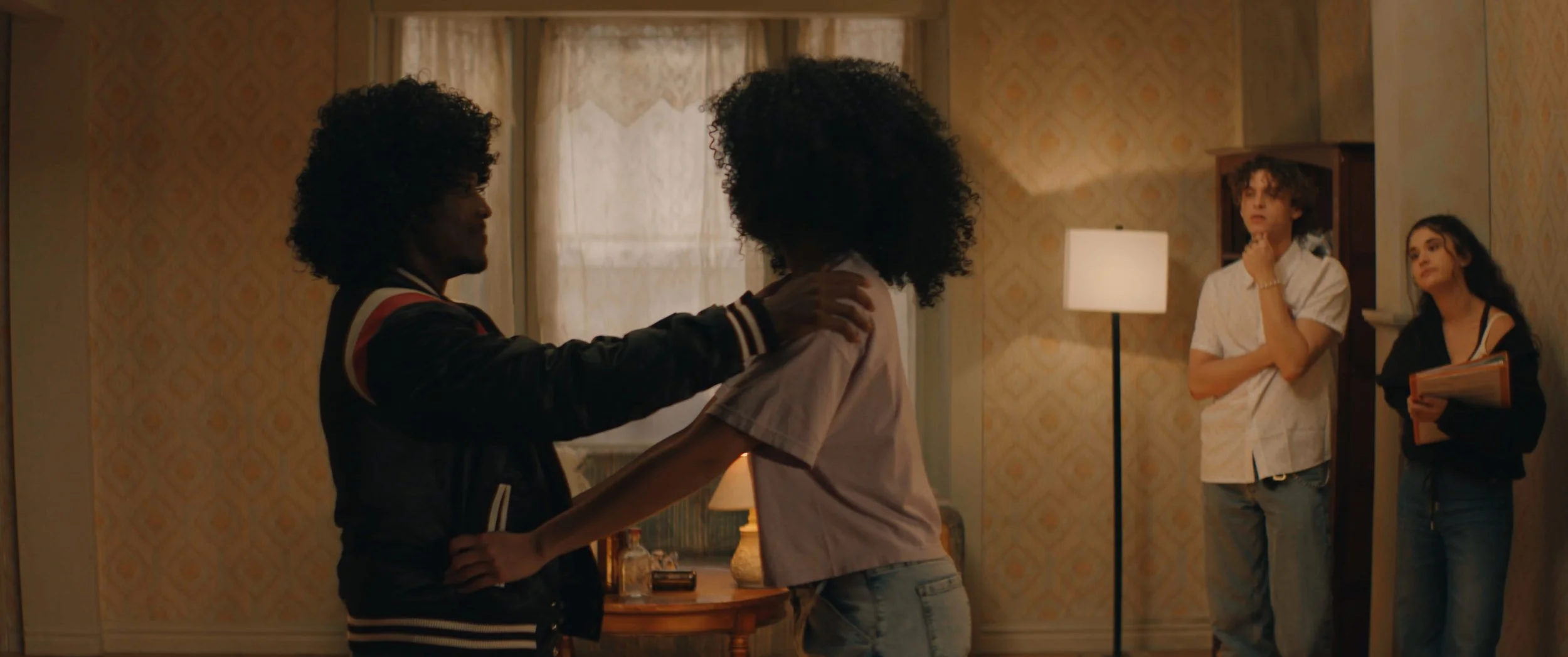 Two people with curly hair are facing each other, with one placing their hand on the other's shoulder in an indoor setting, while two others watch from the background.