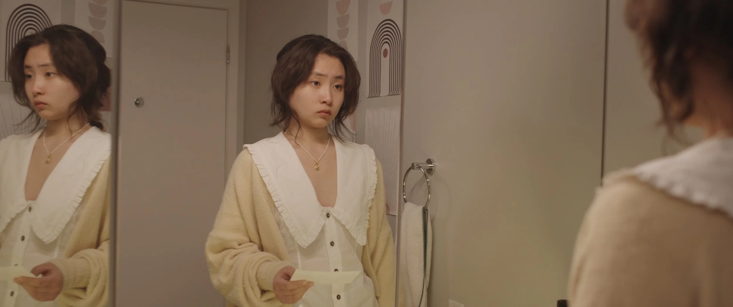 A young woman with short dark hair standing in a bathroom, looking at her reflection in the mirror with a pensive expression. She is holding a piece of paper and wearing a gold necklace, a beige cardigan, and a white top. The bathroom has a towel ring with a white towel and patterned wall decor.