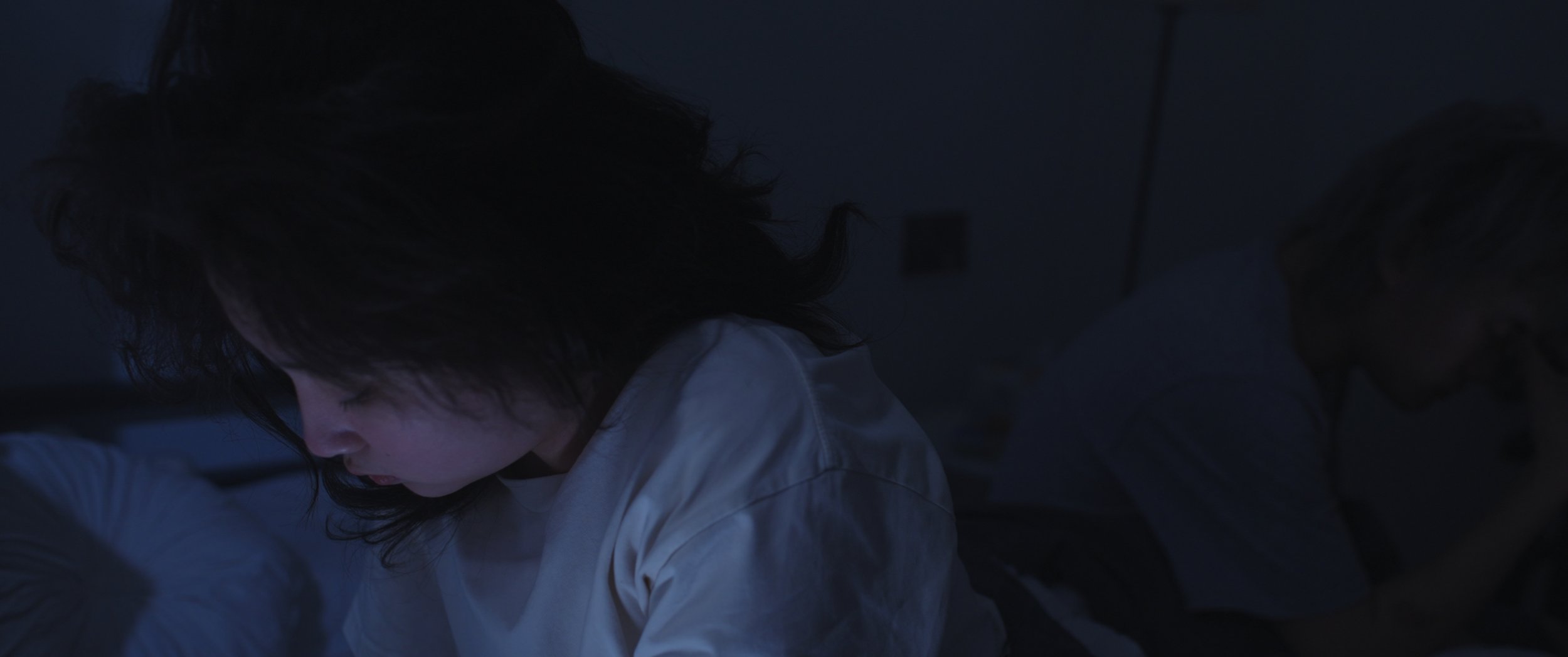 A woman with dark hair wearing a white shirt looking down in a dimly lit room.
