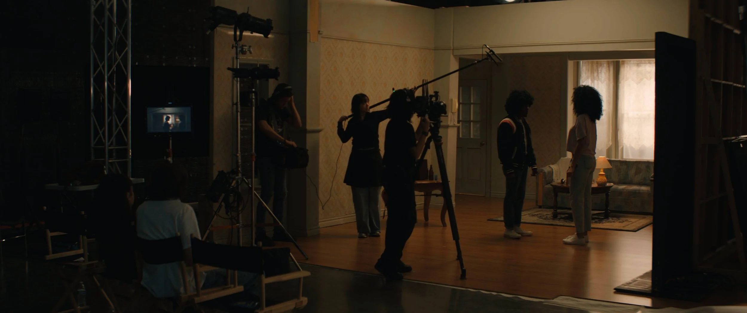 Filming crew captures a scene of two women standing in a room with vintage decor, illuminated by natural light from a window, during a film shoot.