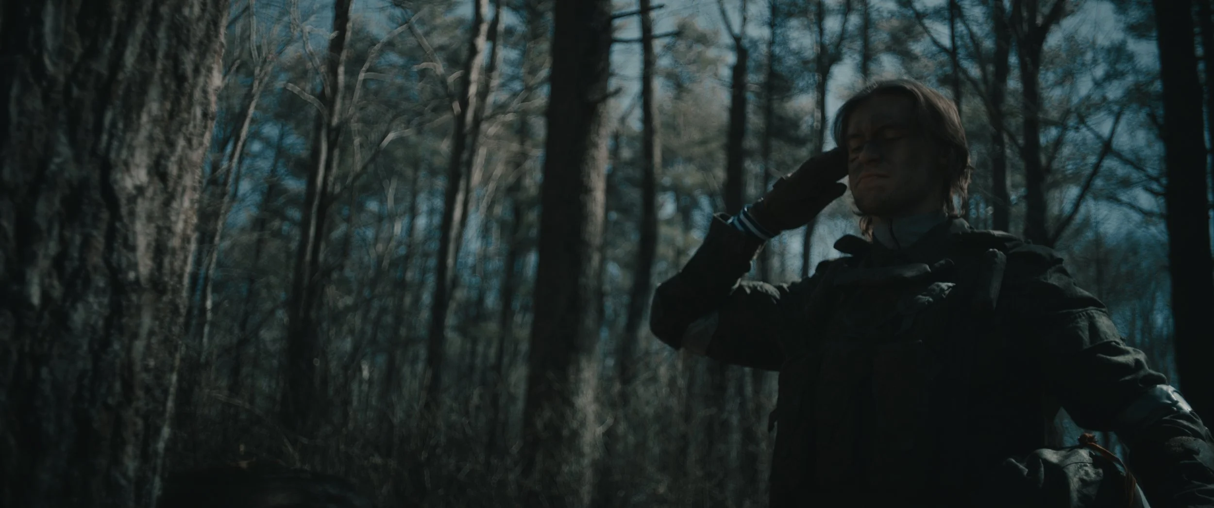 A person wearing glasses and a dark jacket is in a dense forest, with one hand raised to the side of their head, possibly saluting or adjusting their glasses.