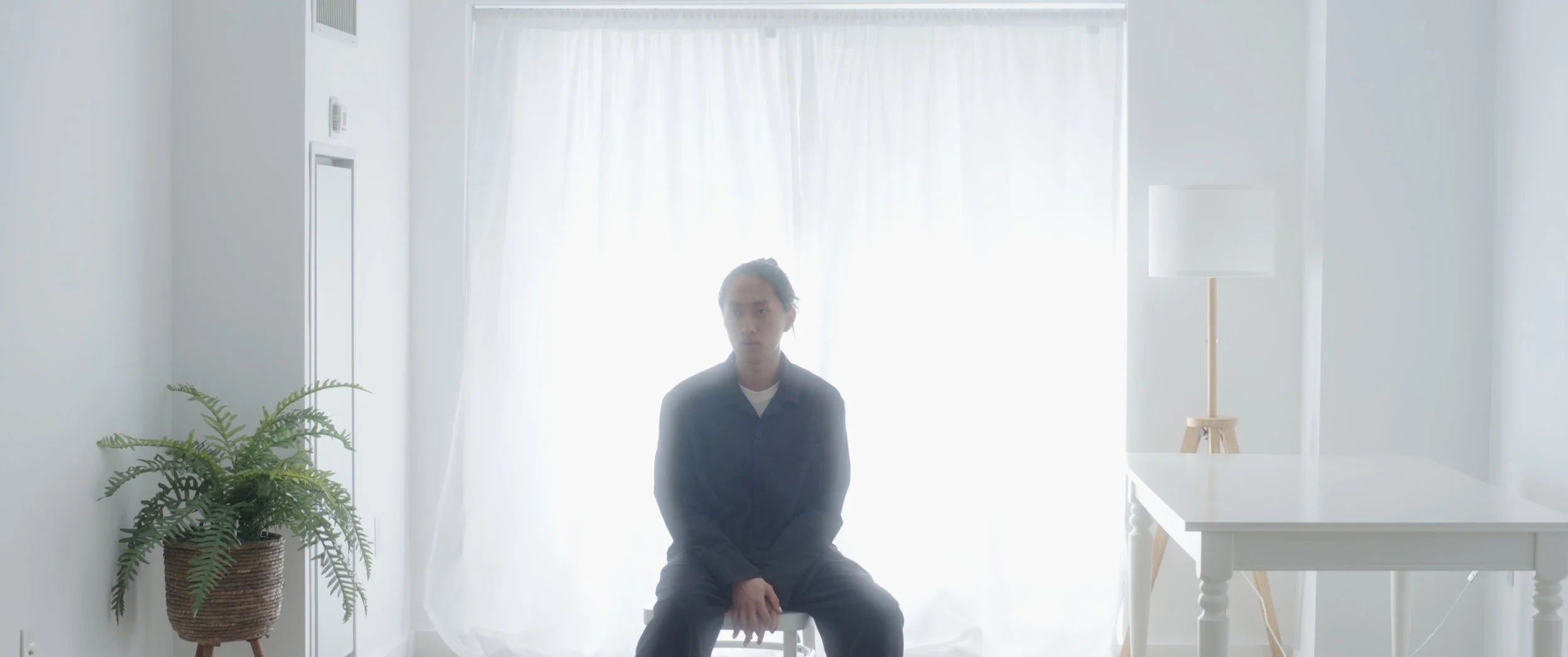A man sitting on a white chair in a bright, minimalist room with white walls, a floor lamp, a potted plant, and sheer white curtains.