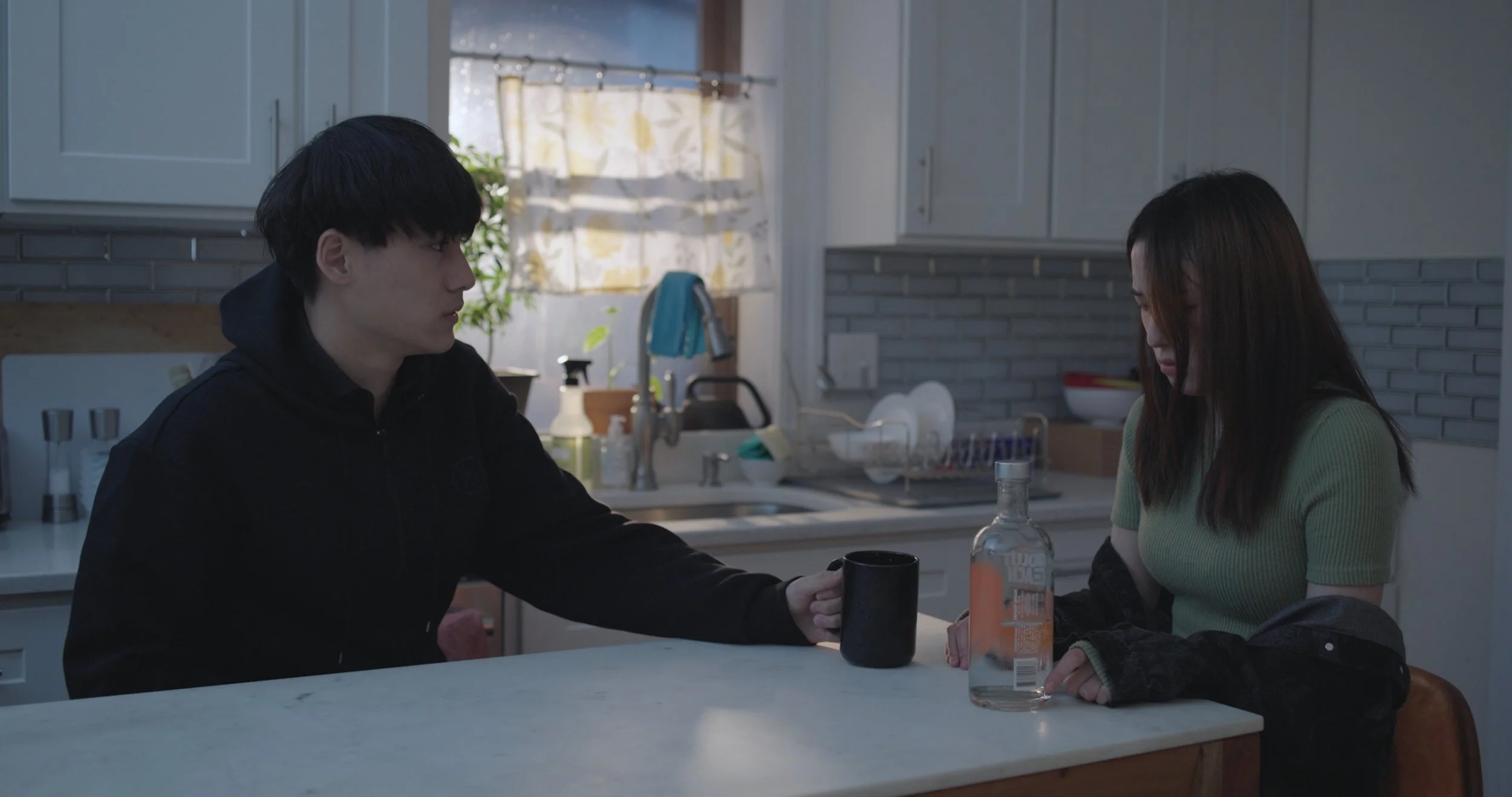 A young man and woman sitting at a kitchen table, with the young man passing a black cup to the woman, holding her hand. The kitchen has white cabinets, a window with a curtain, and various bottles on the counter.