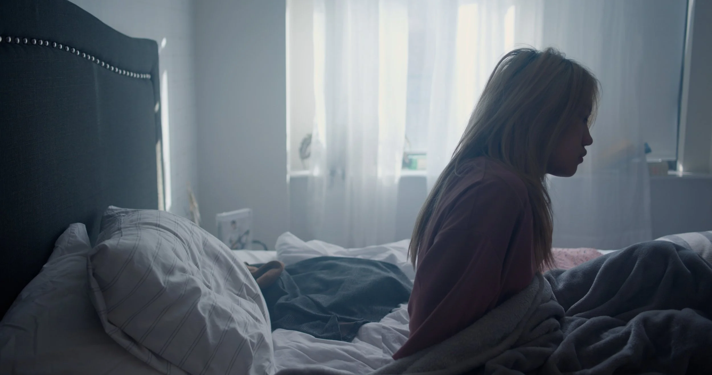 A woman with blonde hair sitting on the edge of a bed, looking down, in a softly lit bedroom with white curtains and a headboard with decorative studs.