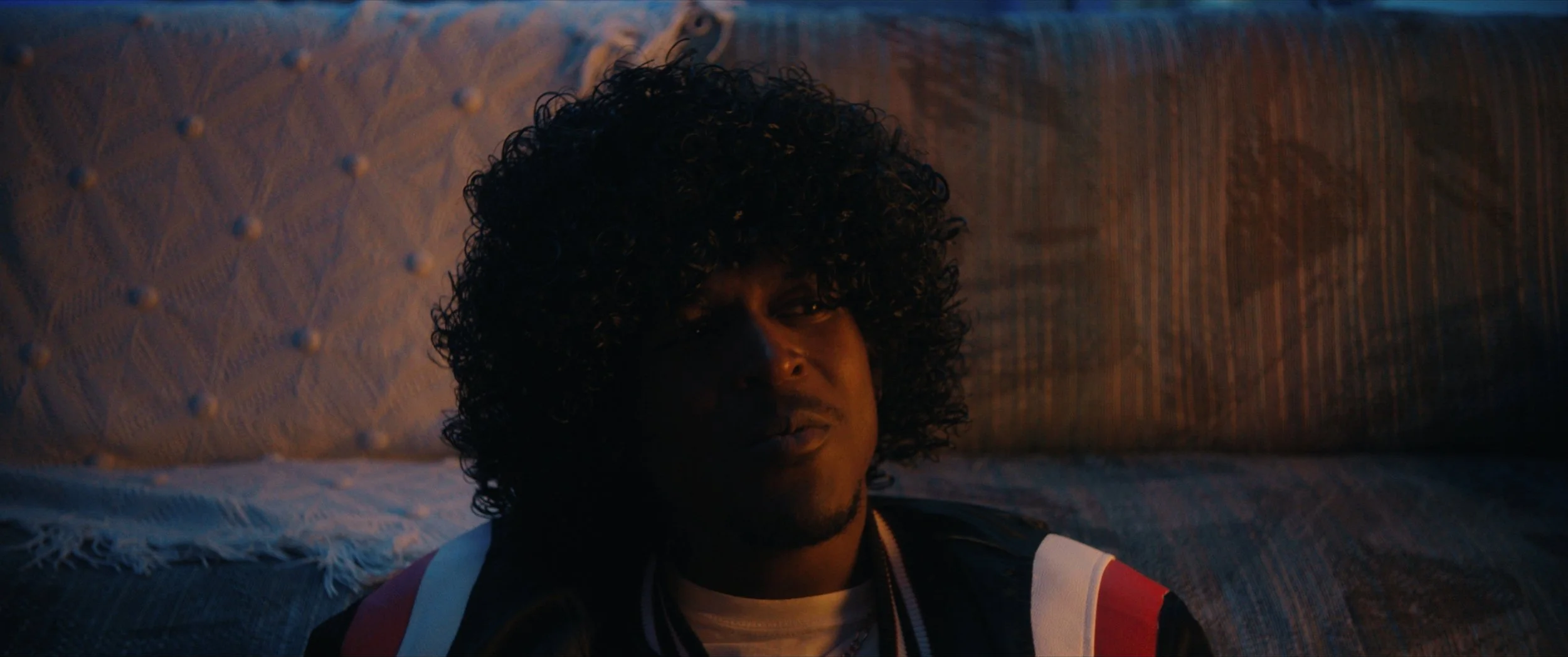 A person with curly hair resting on a quilted bed with a textured pillow in warm lighting.