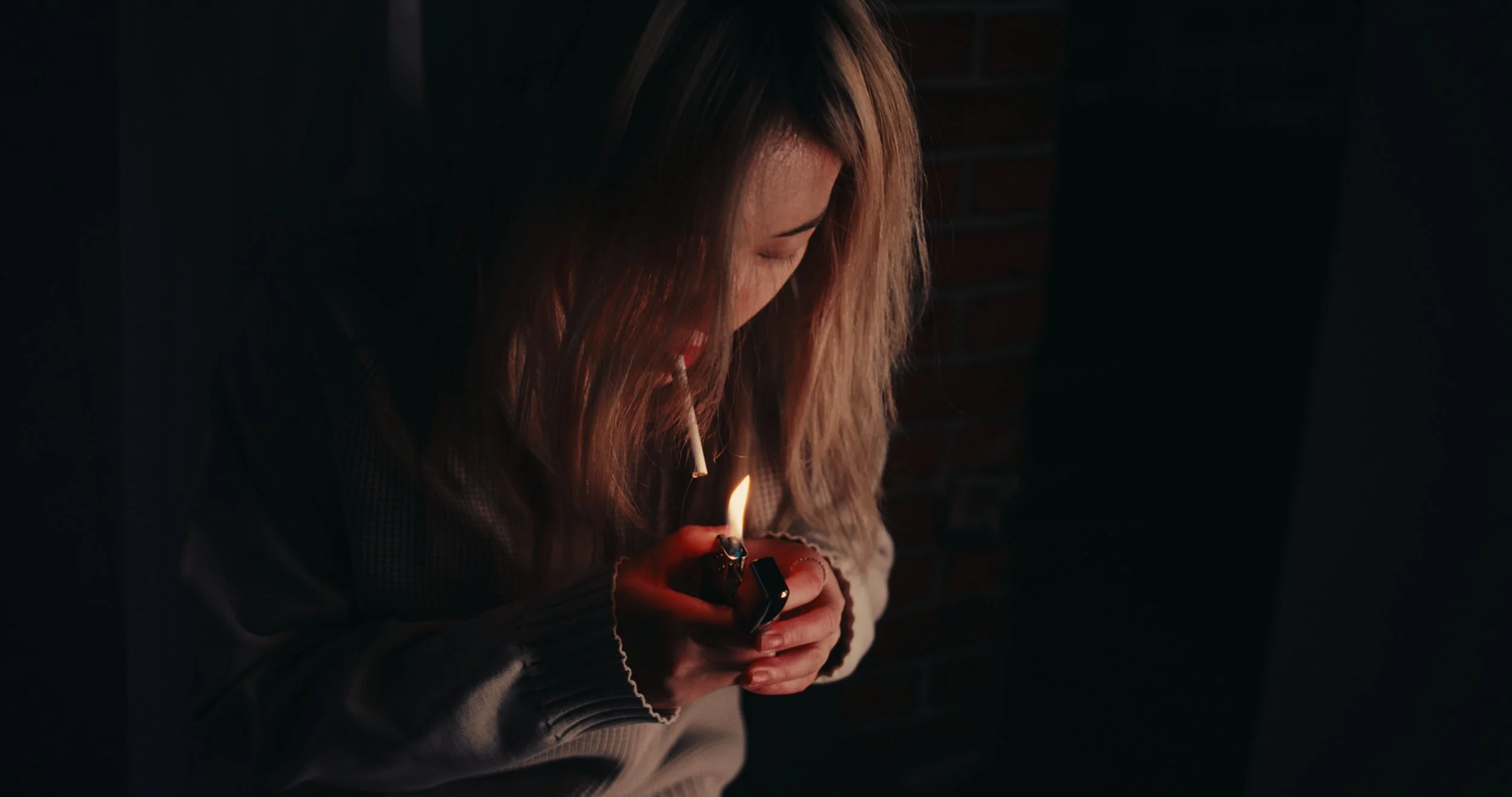 Person with long hair lighting a cigarette in dark setting.