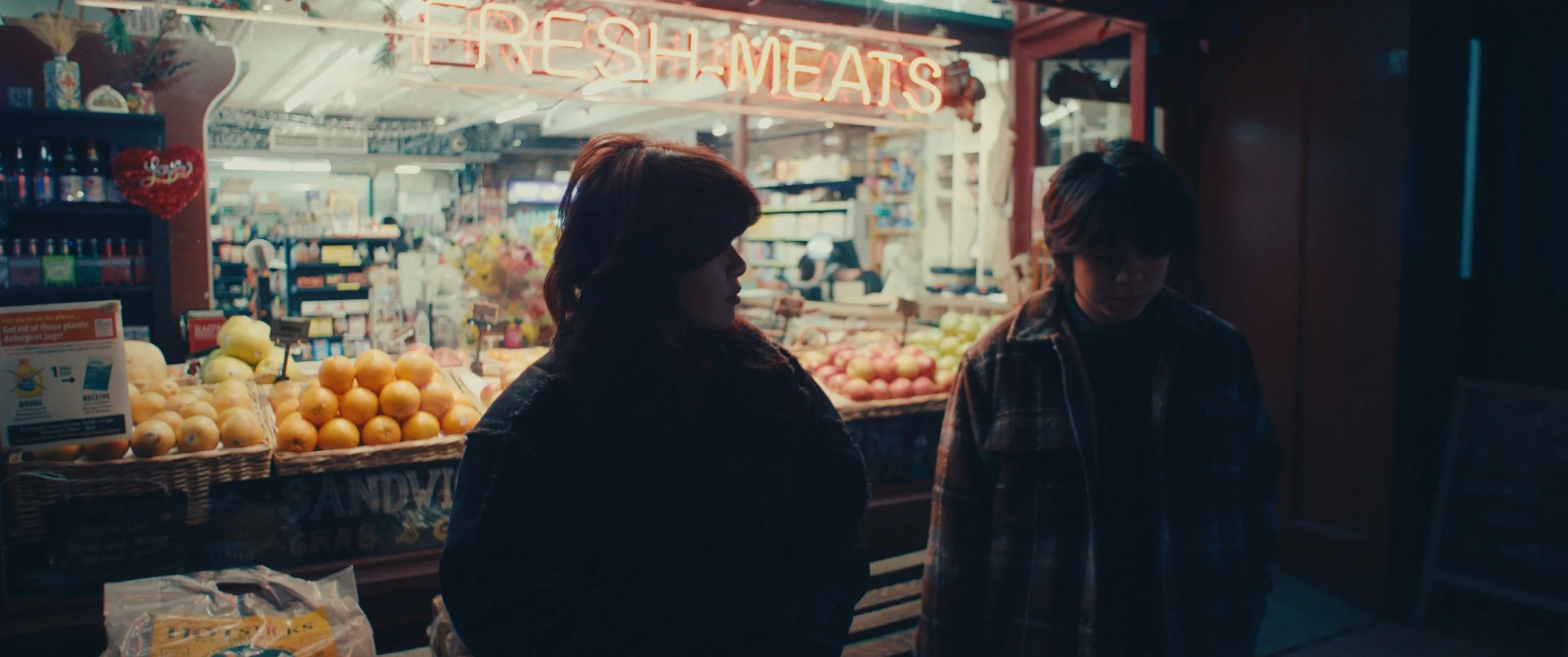 A neon sign reading 'FRESH MEATS' illuminates the entrance of a grocery store. Inside, two people, a woman and a man, stand near the produce section with baskets of oranges, apples, and other fruits. The scene is dimly lit with a moody, cinematic ambiance.