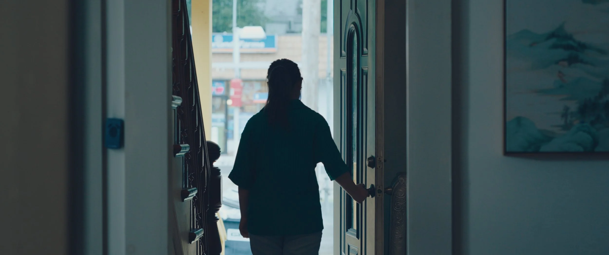 Person opening a door to the outside, seen from behind, in a hallway with artwork on the walls.