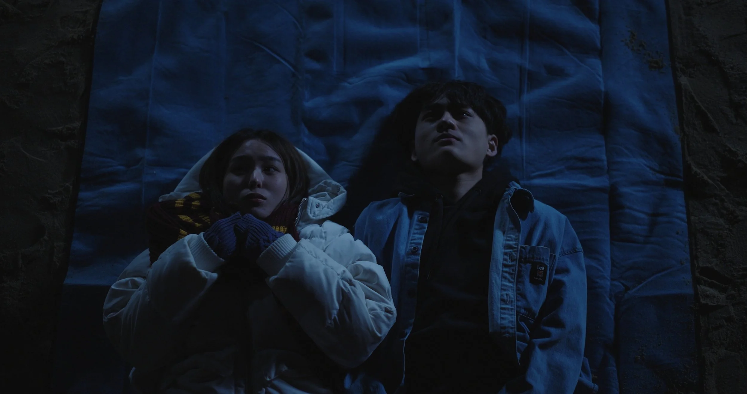 Two young people lying on a blue sleeping bag, looking up at the dark sky, dressed in warm winter clothing with a dark rocky background.