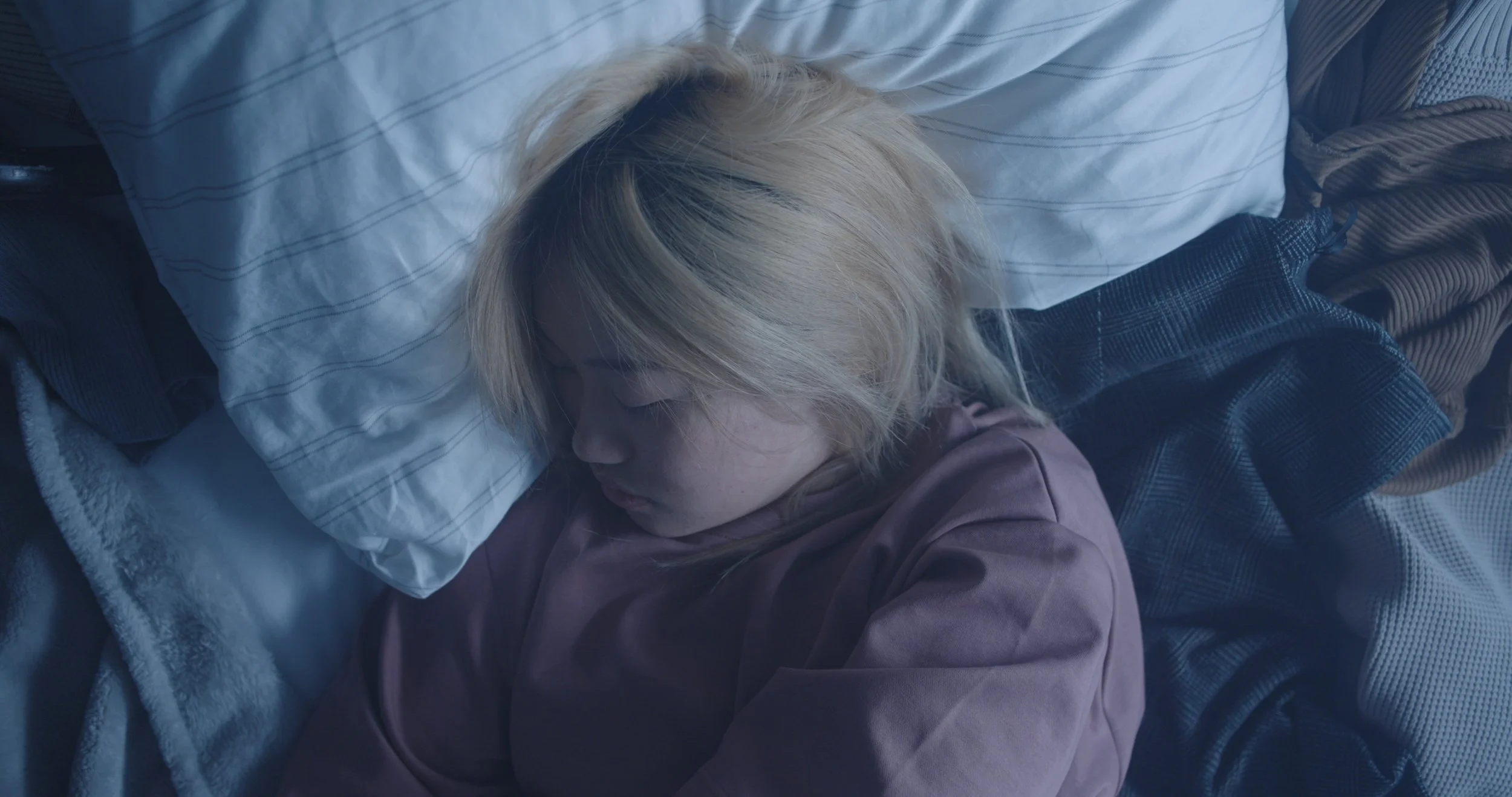 A person with blonde hair sleeping on a bed with a white pillow and layered blankets.